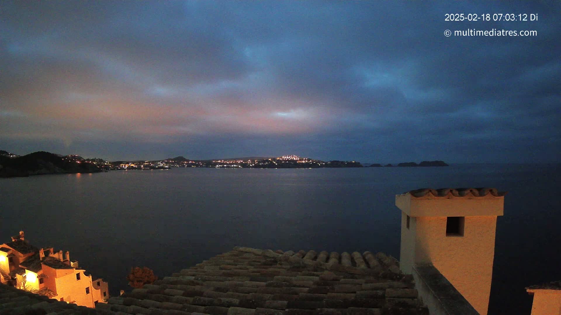 A nighttime view of a calm body of water with a town's lights visible across the bay, under a dark, cloudy sky.