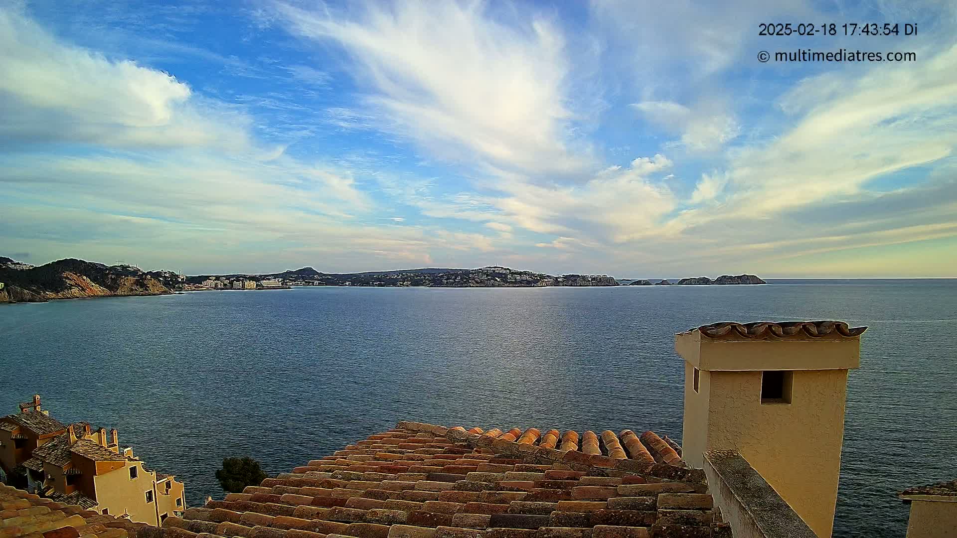 A tranquil coastal scene under a partly cloudy sky shows a calm sea stretching to a distant shoreline with buildings, viewed from a rooftop with terracotta tiles and a stucco chimney.