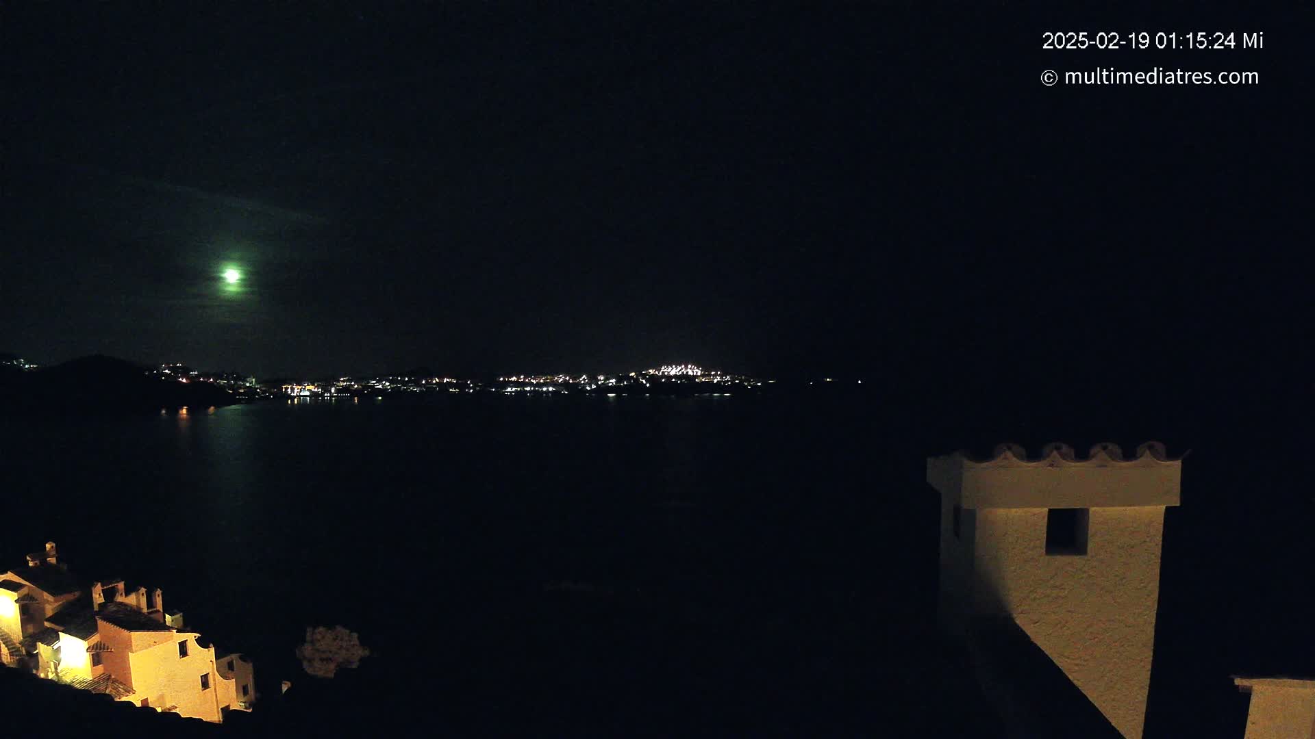 A nighttime view of a moonlit body of water, with a distant town's lights visible across the water and illuminated buildings in the foreground.