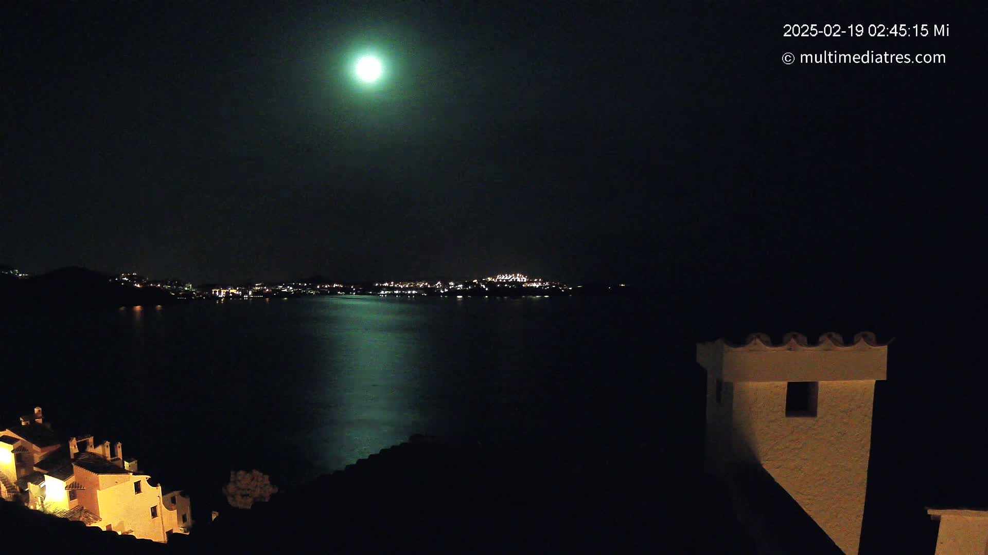 A bright moon illuminates a nighttime scene of a dark body of water reflecting the lights of a distant town, with illuminated buildings visible in the foreground.