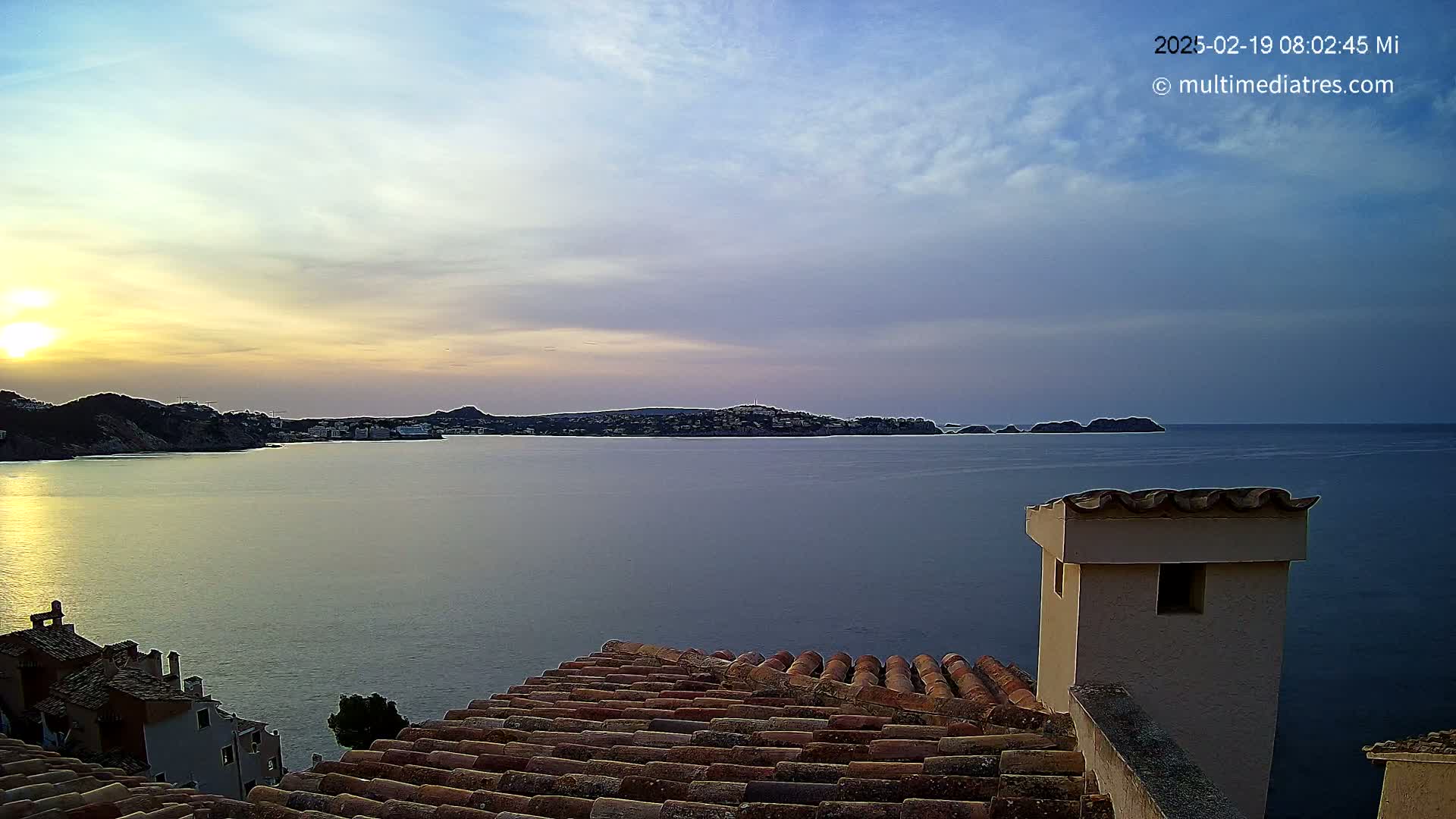 A tranquil sunset over a calm sea, viewed from a tiled rooftop with a chimney, shows a distant coastline.
