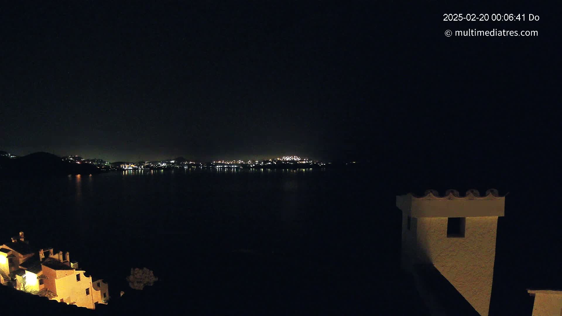A nighttime view of a city's lights across a dark body of water, with some illuminated buildings in the foreground.