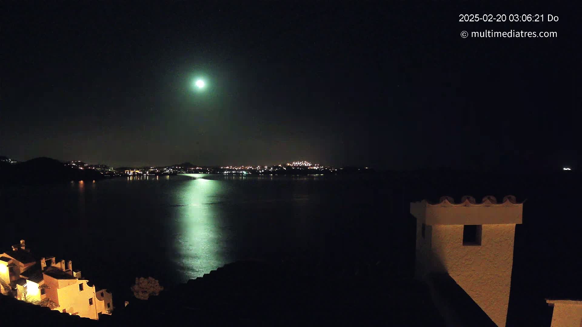 A nighttime view of a moonlit body of water reflecting city lights across from a hillside village under a clear sky.