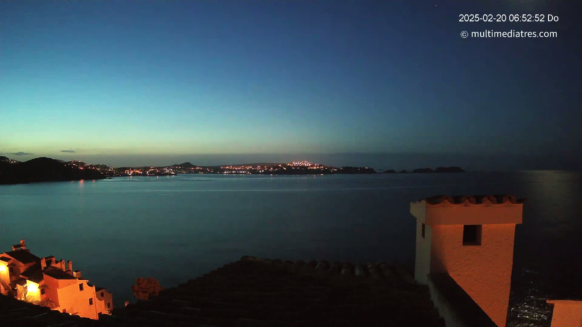 A nighttime coastal scene shows a calm body of water separating a dimly lit town from an orange-lit building perched on a cliff overlooking the water.