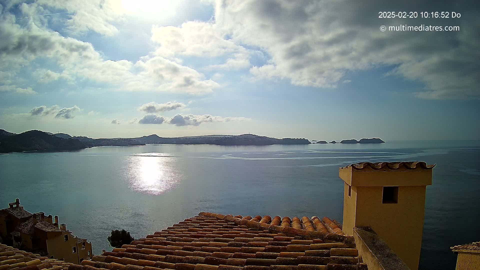 A partly sunny day shows a calm sea reflecting sunlight, with distant hills and islands under a mostly cloudy sky, viewed from a tiled rooftop with a chimney.
