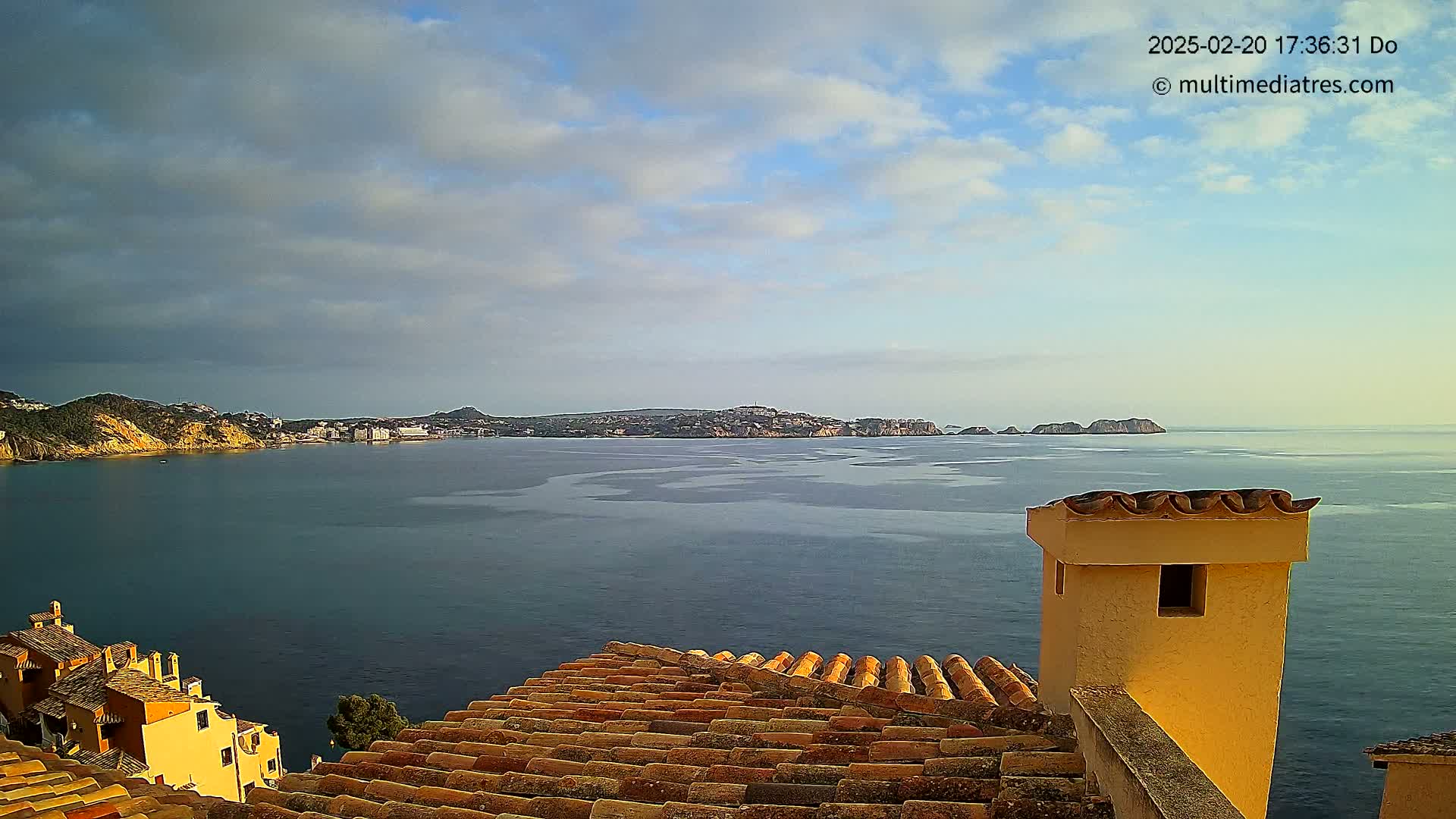 A partly cloudy sky overlooks a calm sea with a distant town and islands visible from a terracotta roof.