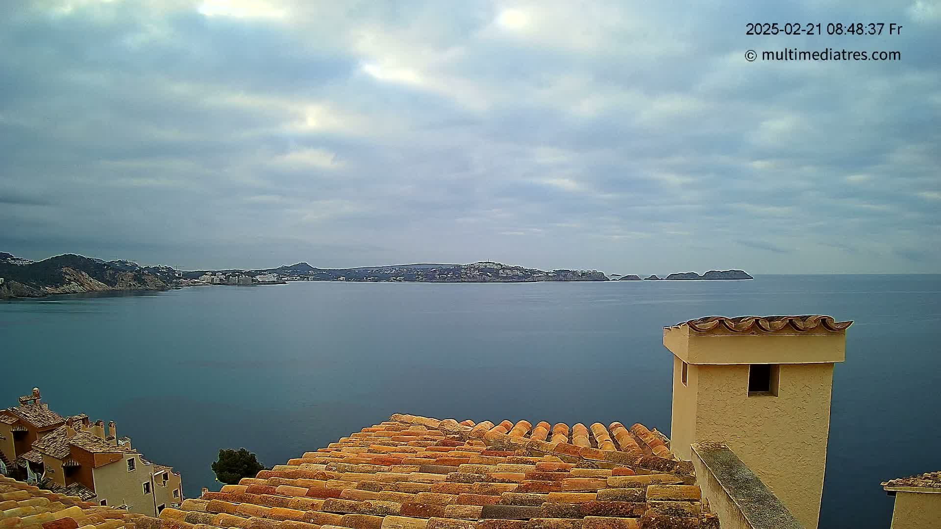 A calm, overcast day reveals a coastal view of a town with terracotta rooftops overlooking a tranquil sea and distant islands.