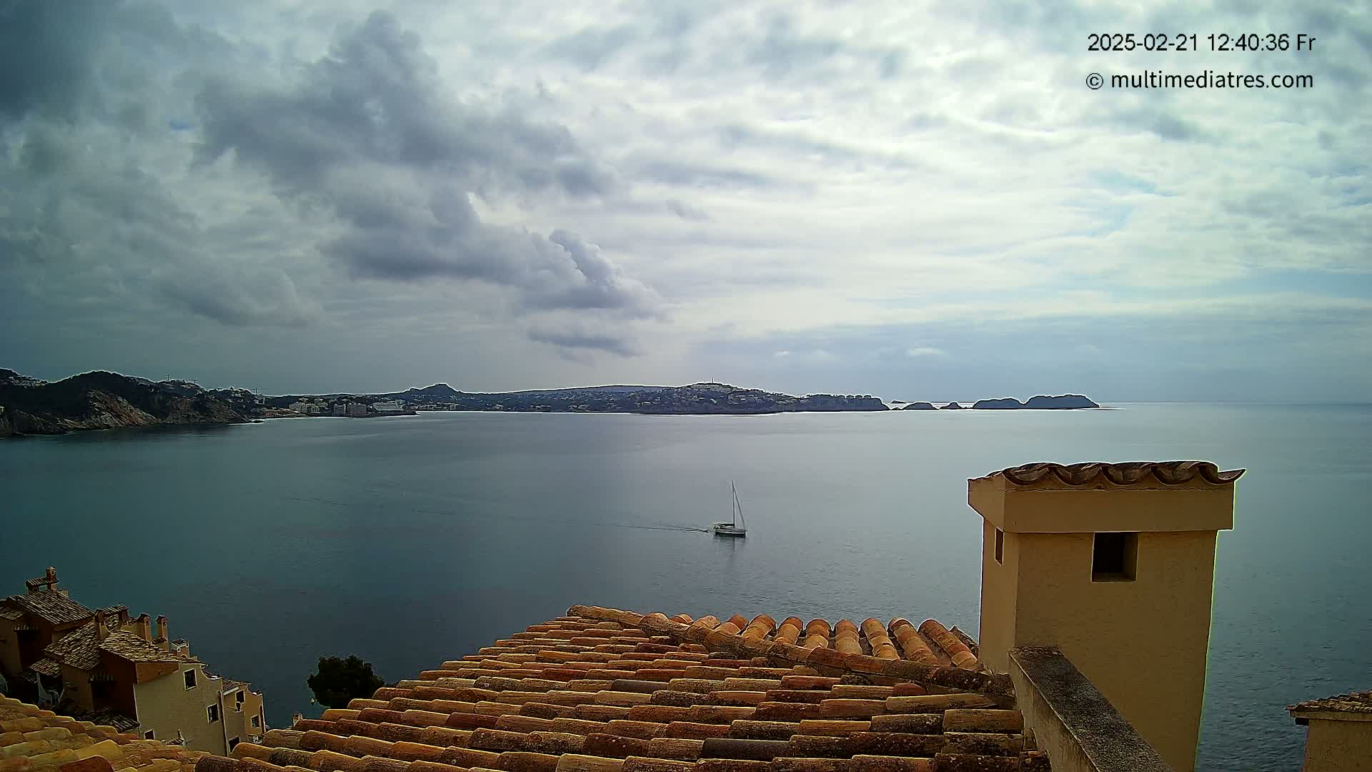 A sailboat is visible on a calm sea under a cloudy sky, viewed from a tiled rooftop overlooking a coastal town and distant islands.
