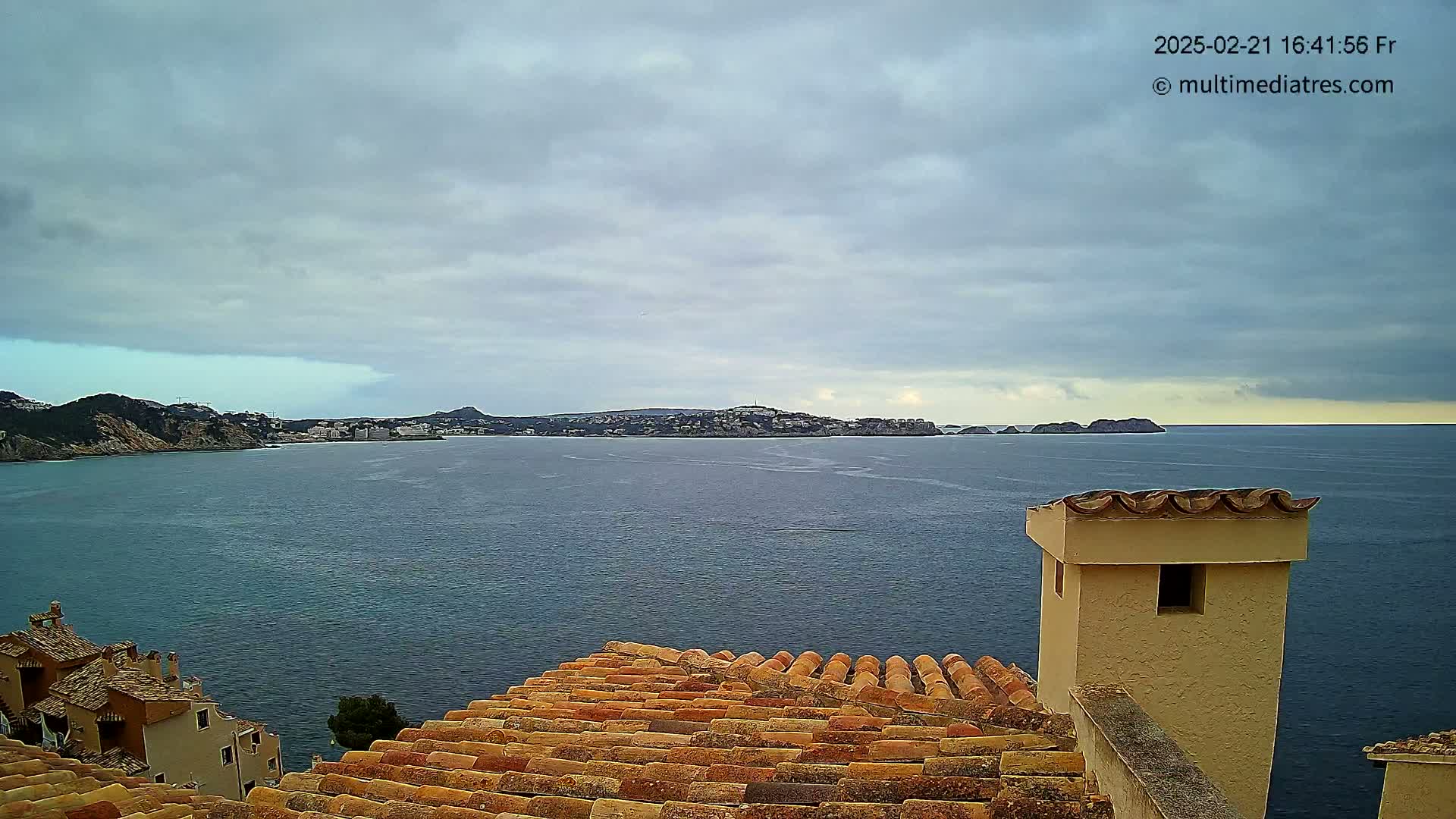A coastal view shows a calm sea under a cloudy sky, with buildings and terracotta rooftops in the foreground and a town across the water.