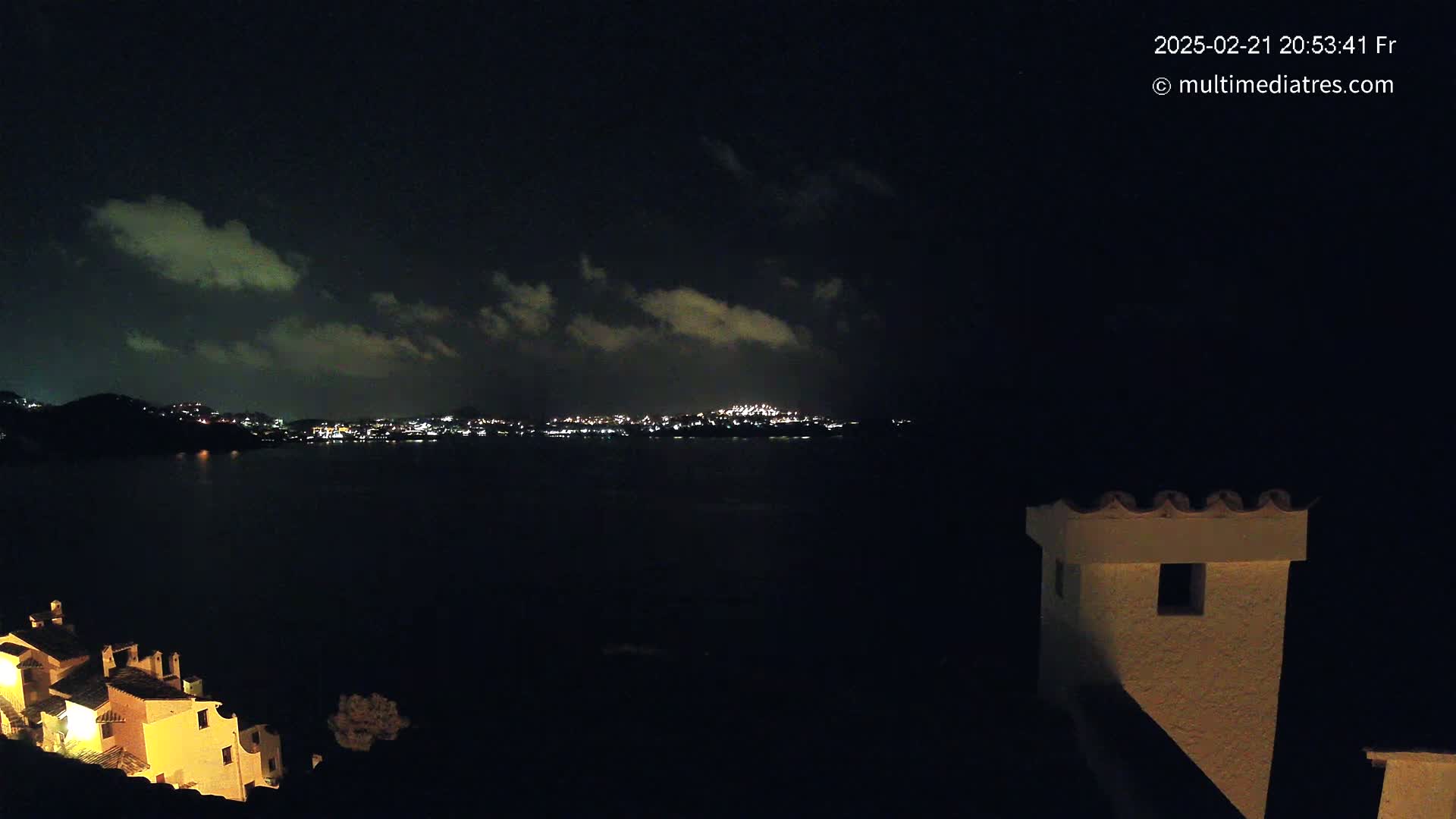 A nighttime view across a dark body of water shows a distant city skyline illuminated with lights under a partly cloudy sky.