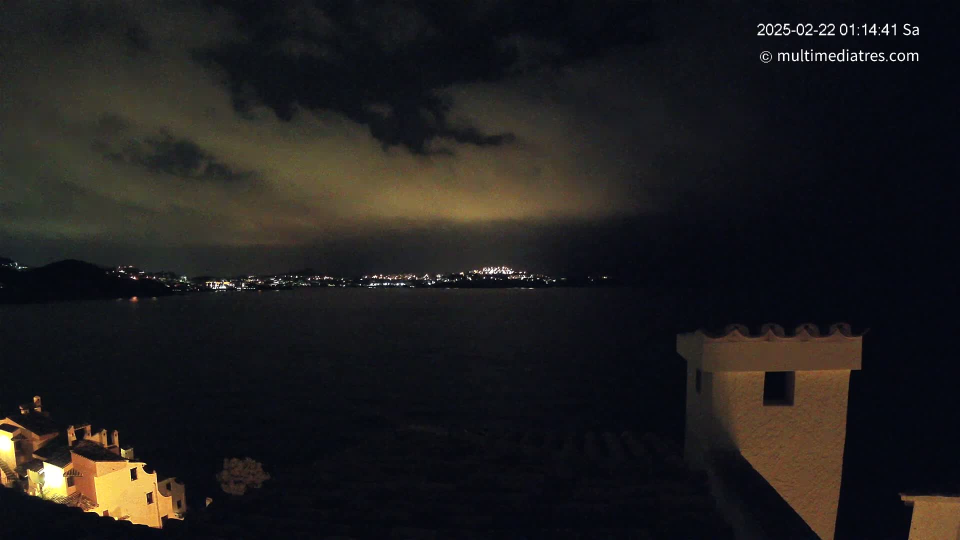 A nighttime view across a dark body of water shows a distant, illuminated city skyline under a cloudy sky, with a portion of a building in the foreground.