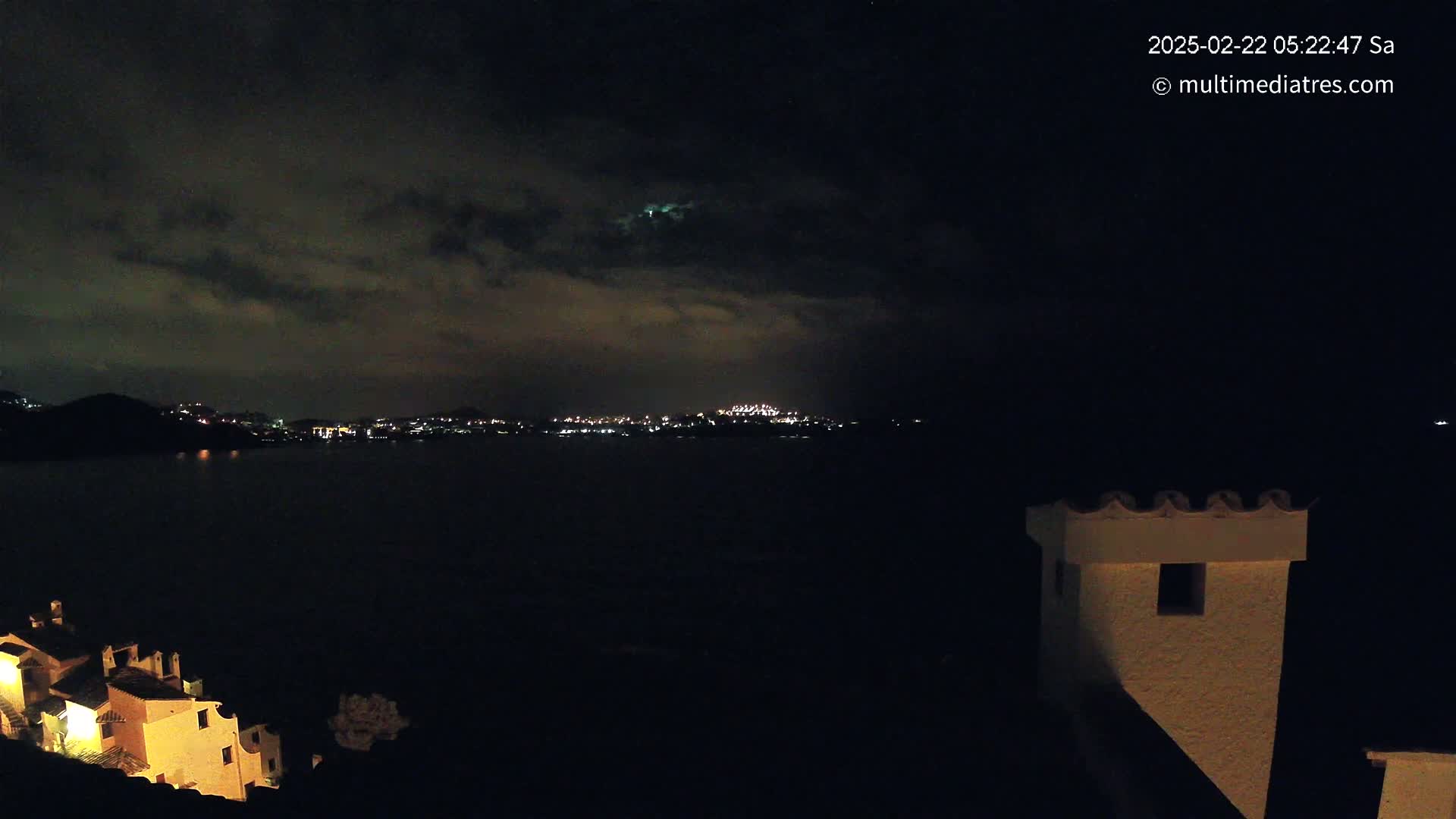 A nighttime view of a town's lights across a dark body of water, under a cloudy sky, with a building partially visible in the foreground.