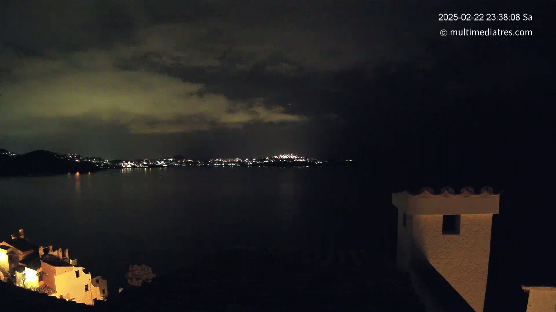 A nighttime view of a city across a dark body of water, under a cloudy sky, with illuminated buildings visible in the foreground.