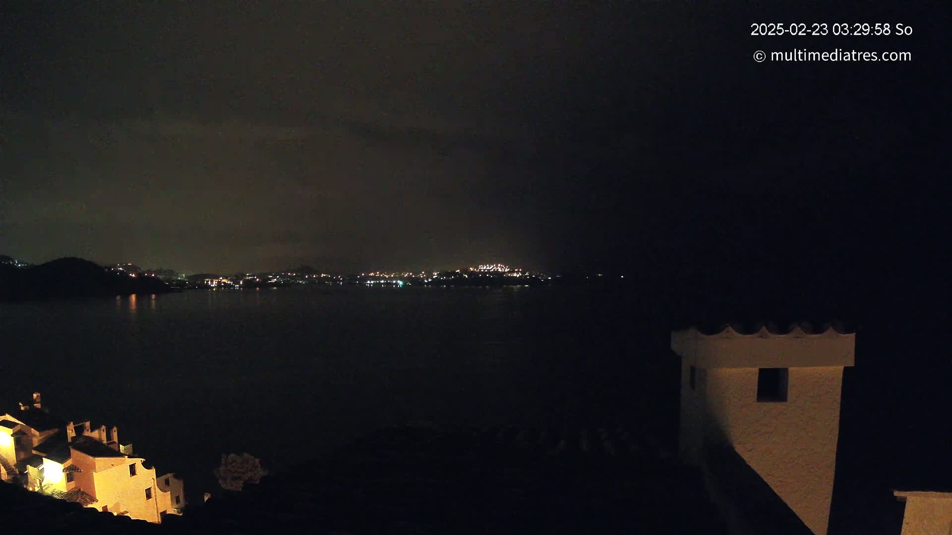 A nighttime view of a dark body of water with a distant city skyline illuminated by lights, seen from a vantage point overlooking buildings with some lights on.