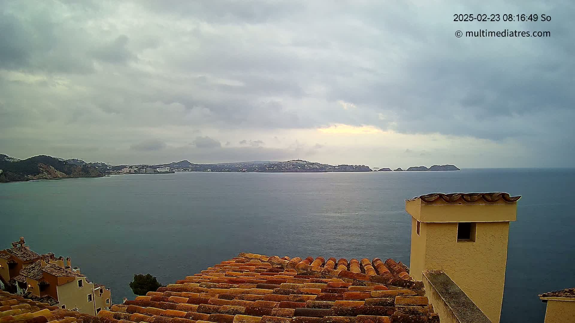 A coastal view under a cloudy sky shows a calm sea separating a town from distant islands and hills, partially obscured by a foreground of orange-tiled rooftops and a tan chimney.