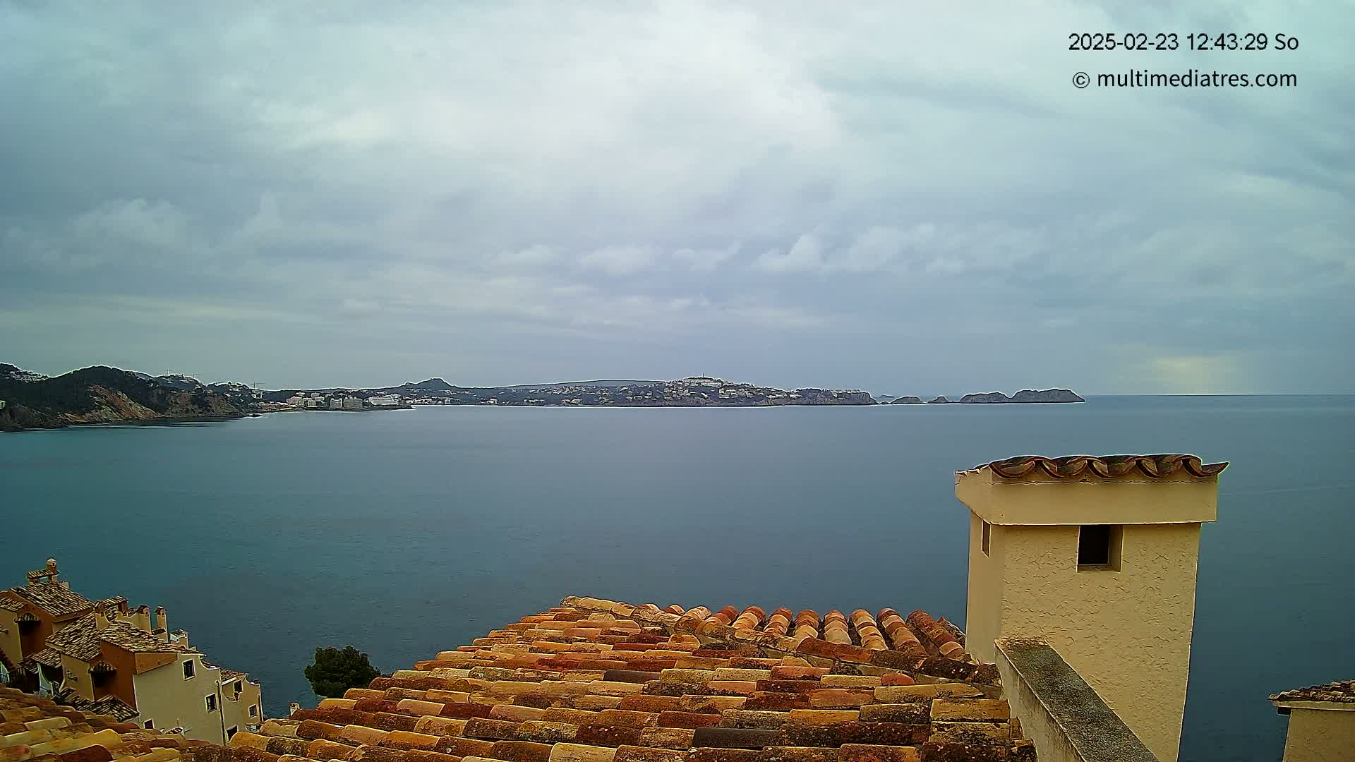 Overlooking a calm sea on an overcast day, a terracotta roof extends toward a distant coastline with buildings and hills.