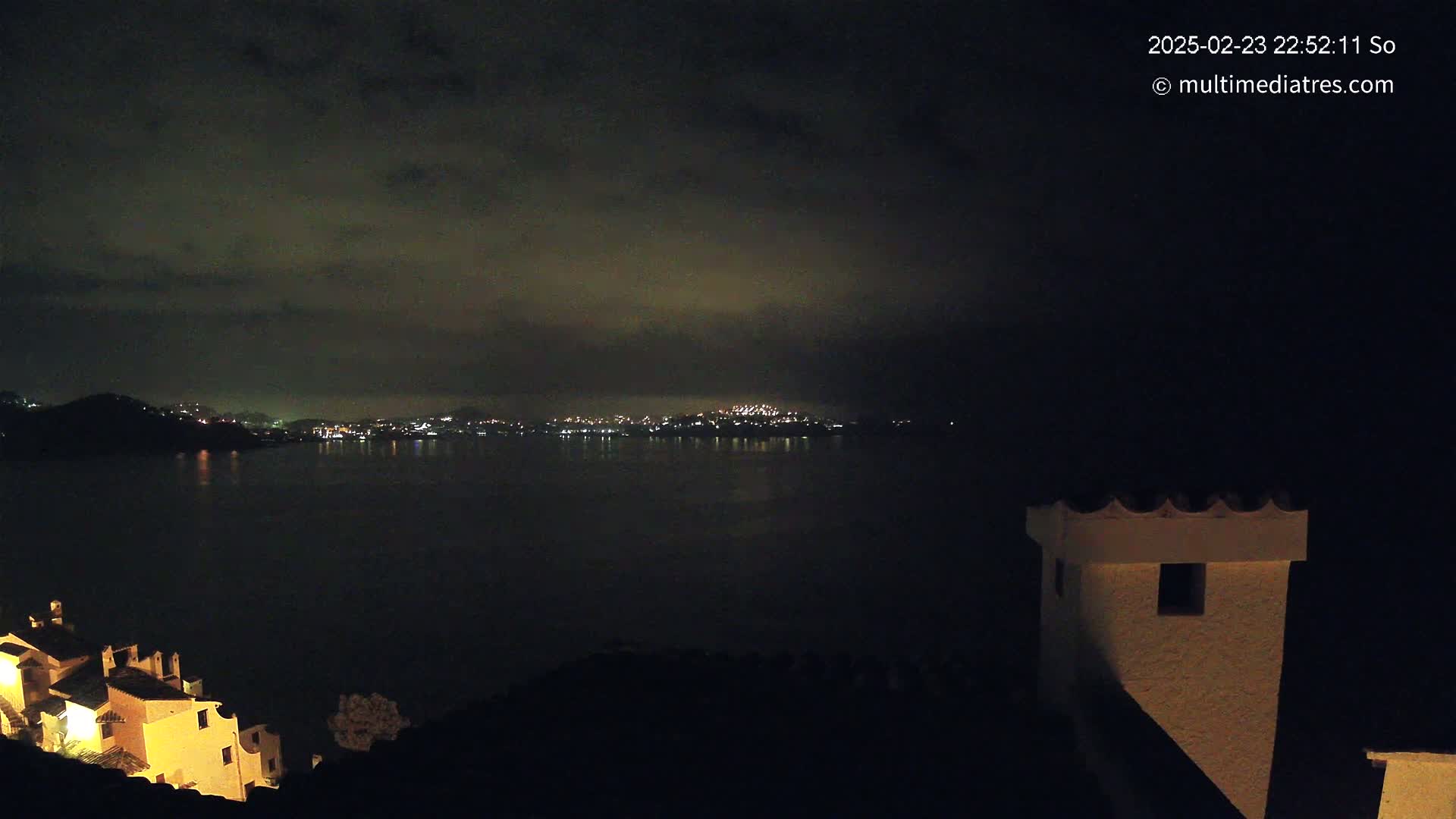 A nighttime view shows a dark body of water reflecting the lights of a city on the opposite shore, with a few illuminated buildings visible in the foreground under a cloudy sky.