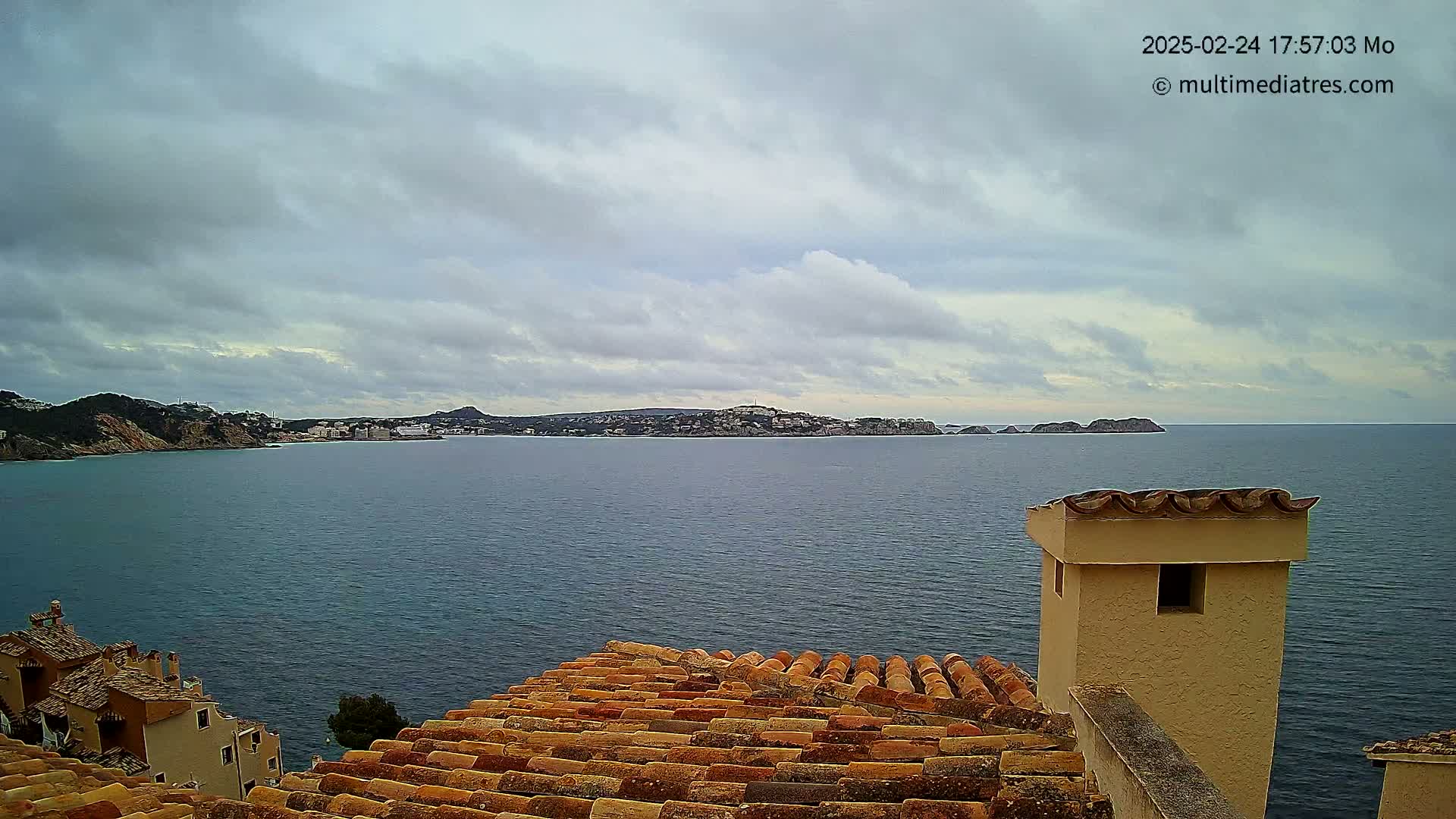 A view from a terracotta roof shows a calm, overcast sea with distant hills and buildings under a cloudy sky.
