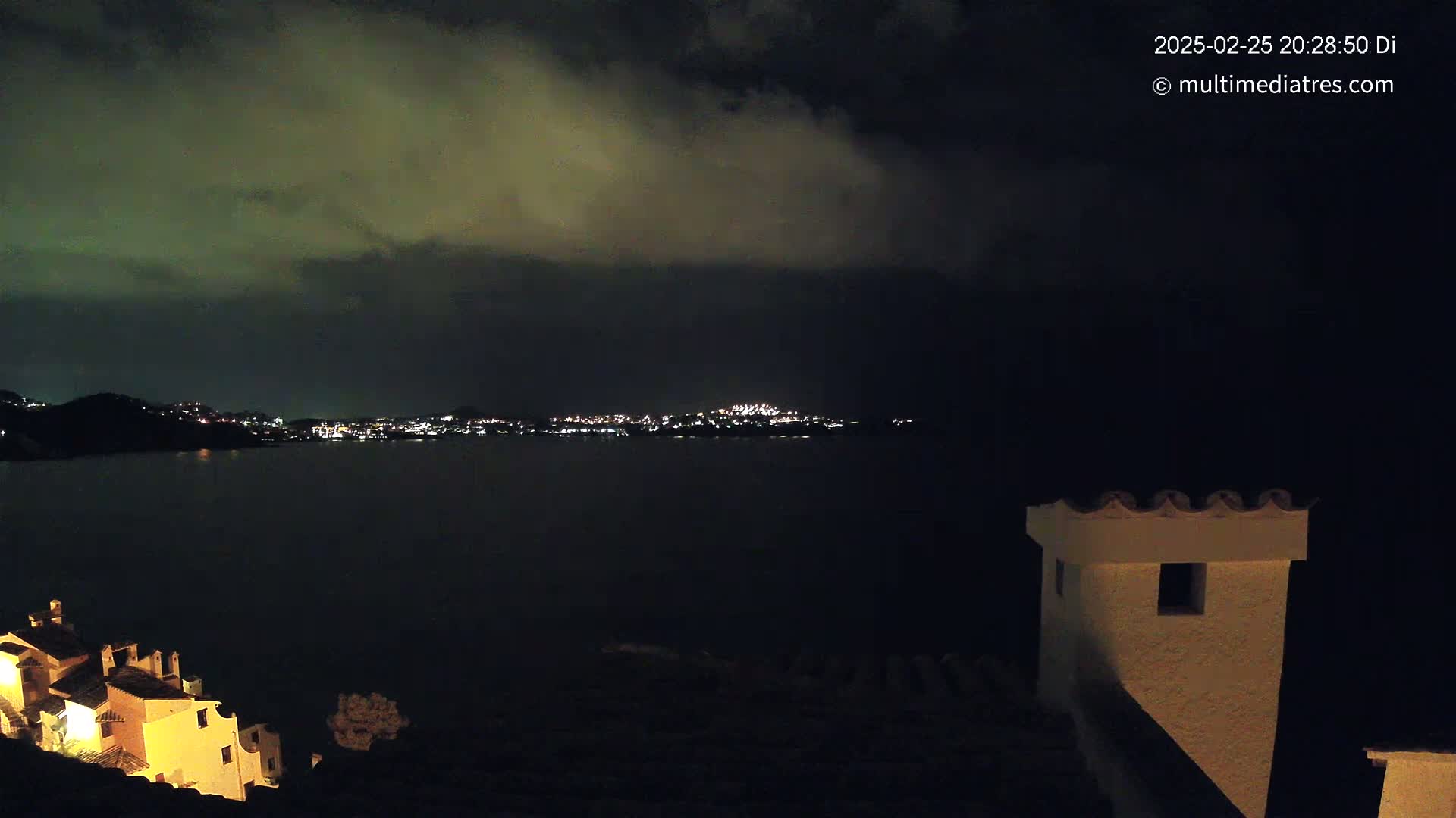 A nighttime view of a dark body of water with a city skyline illuminated by lights across the water, under a cloudy sky, with a partially lit building in the foreground.