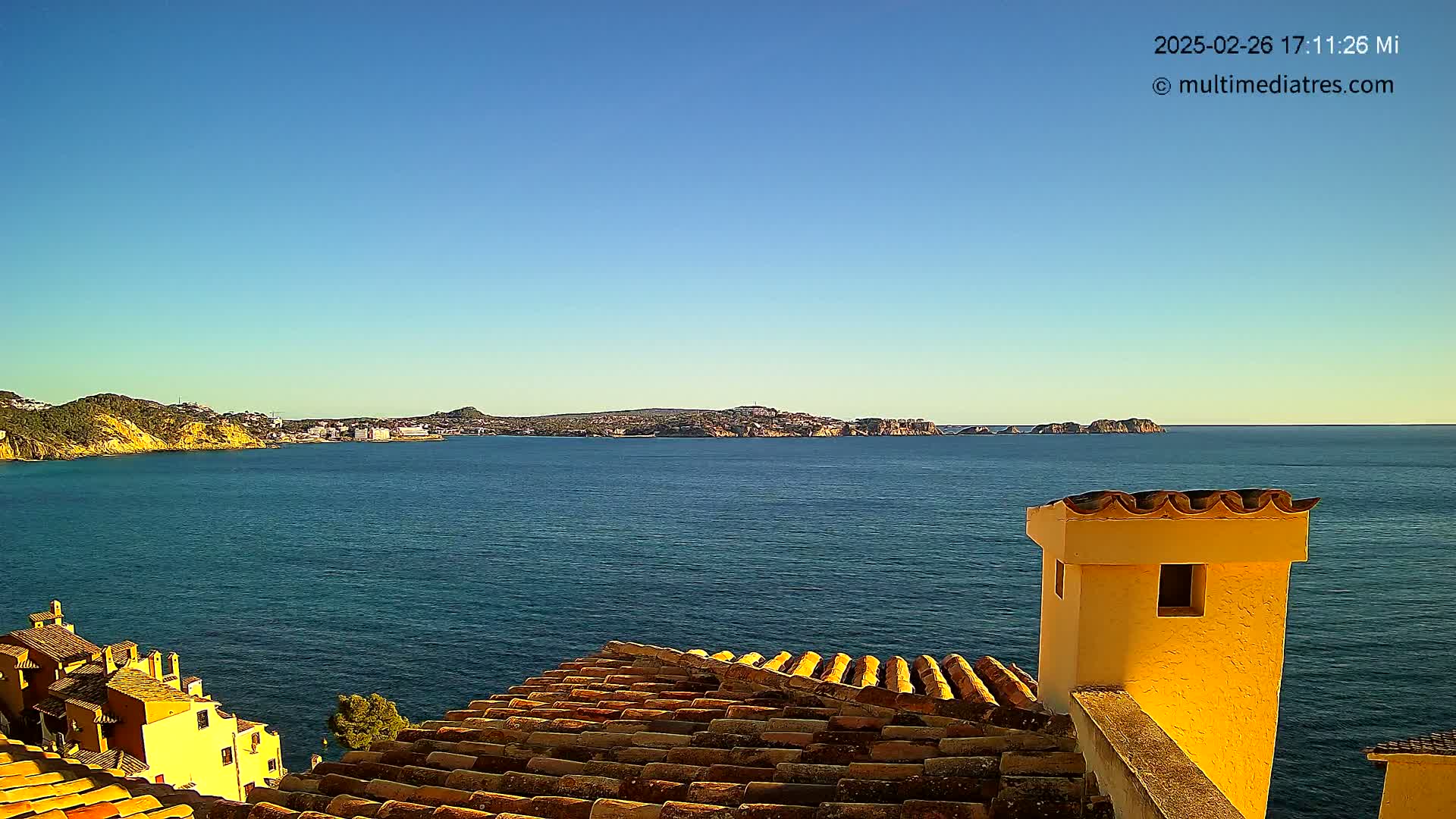 A coastal view on a clear day shows a calm blue sea extending to distant hills and a town, seen from atop a building with a yellow chimney and terracotta roof tiles.