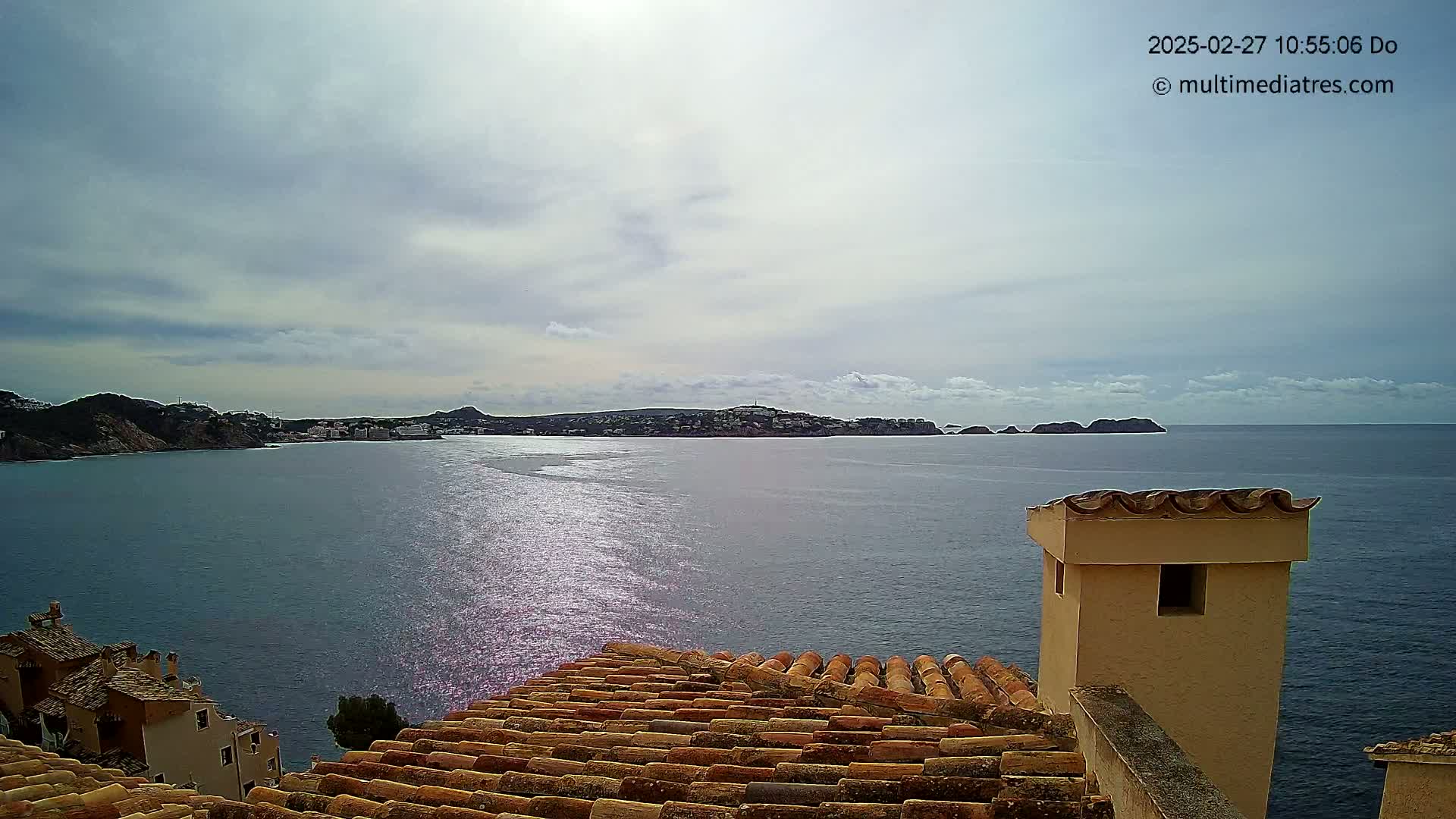 A partly cloudy sky overlooks a calm sea with a coastal town and hills in the distance, viewed from a tiled rooftop with a small building structure.