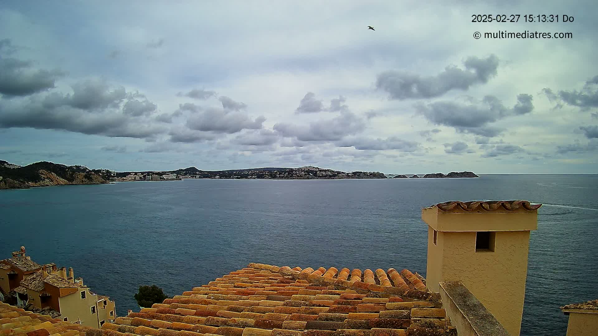 A coastal view of a town under a cloudy sky shows a calm body of water with a distant shoreline, seen from a rooftop with terracotta tiles and a tan chimney.
