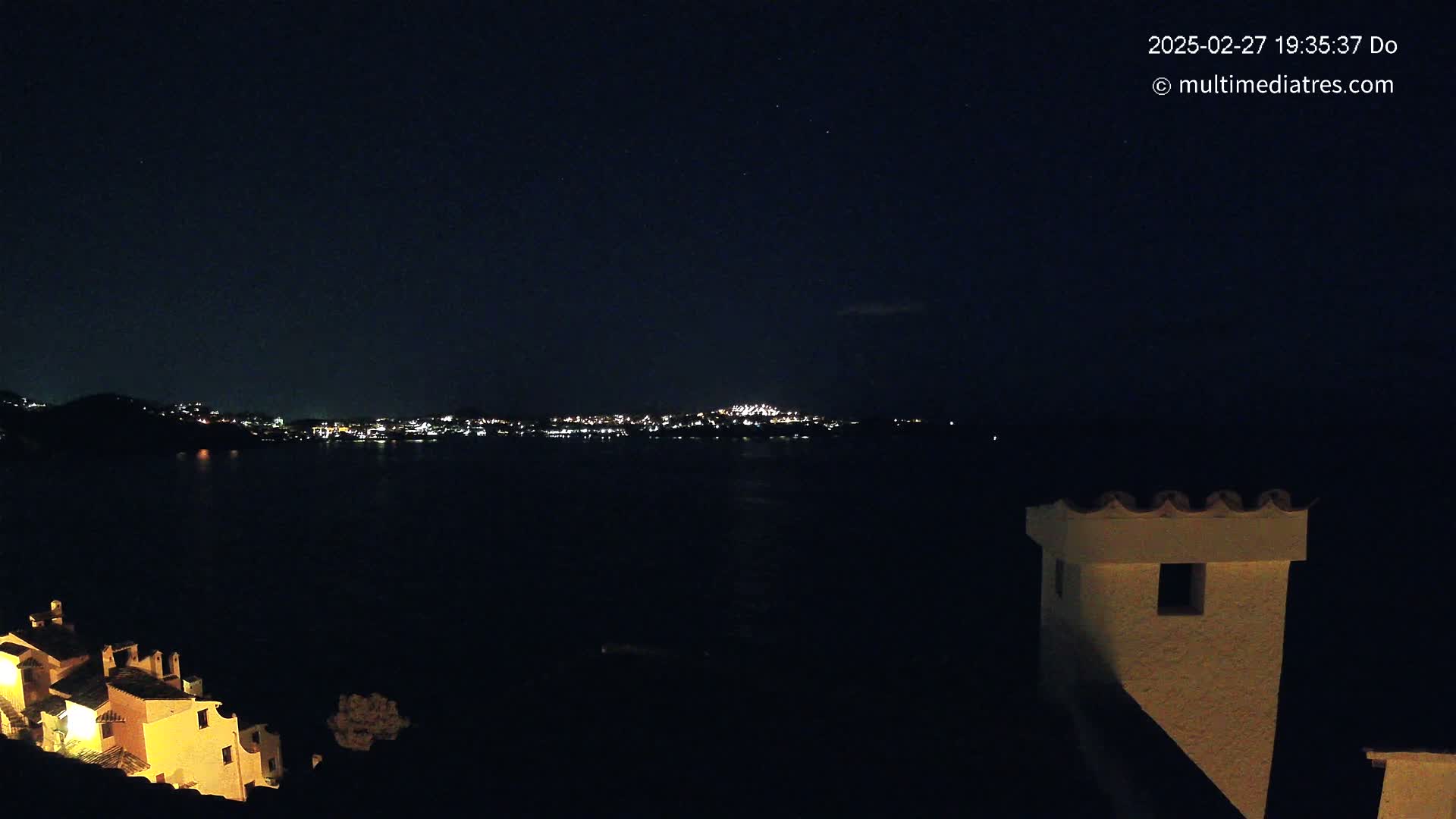 A nighttime view of a city's lights across a dark body of water, with a small illuminated building in the foreground under a clear, starry sky.