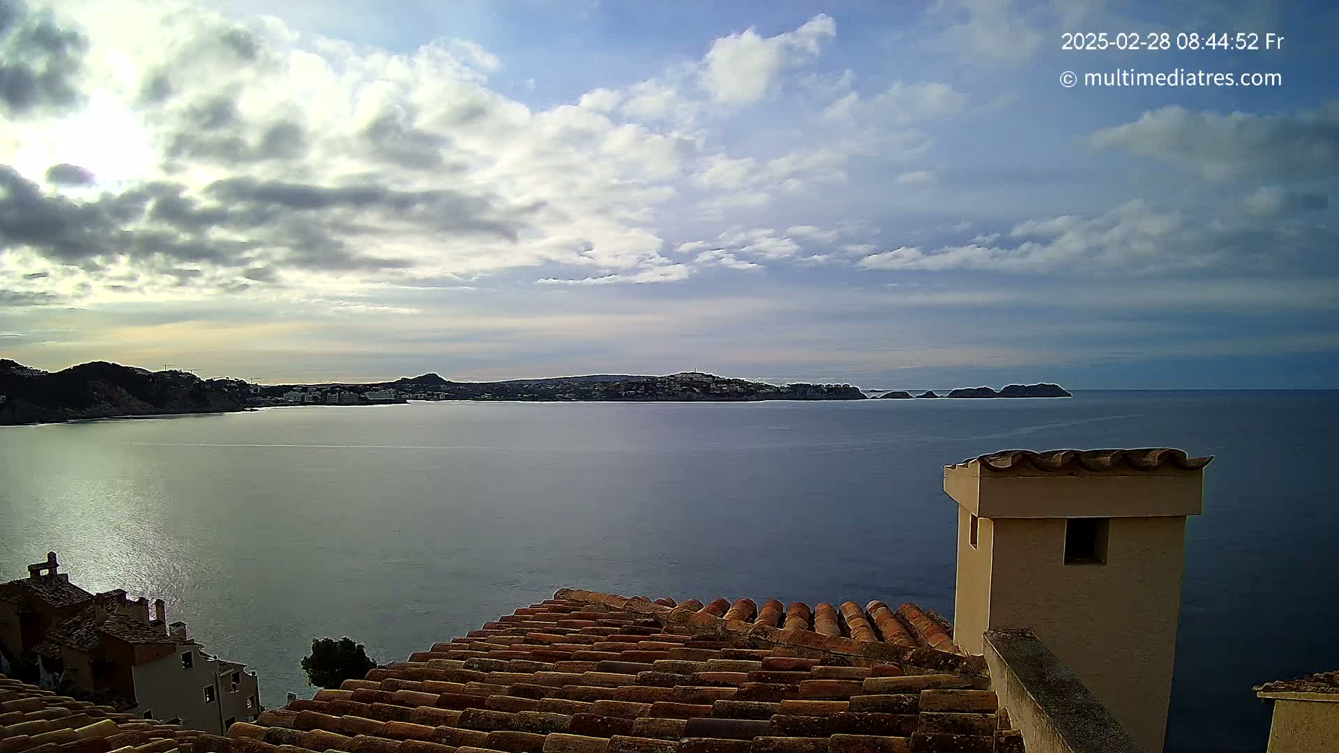 A partly cloudy sky overlooks a calm sea with a distant town nestled between hills, viewed from a terracotta roof with a chimney.