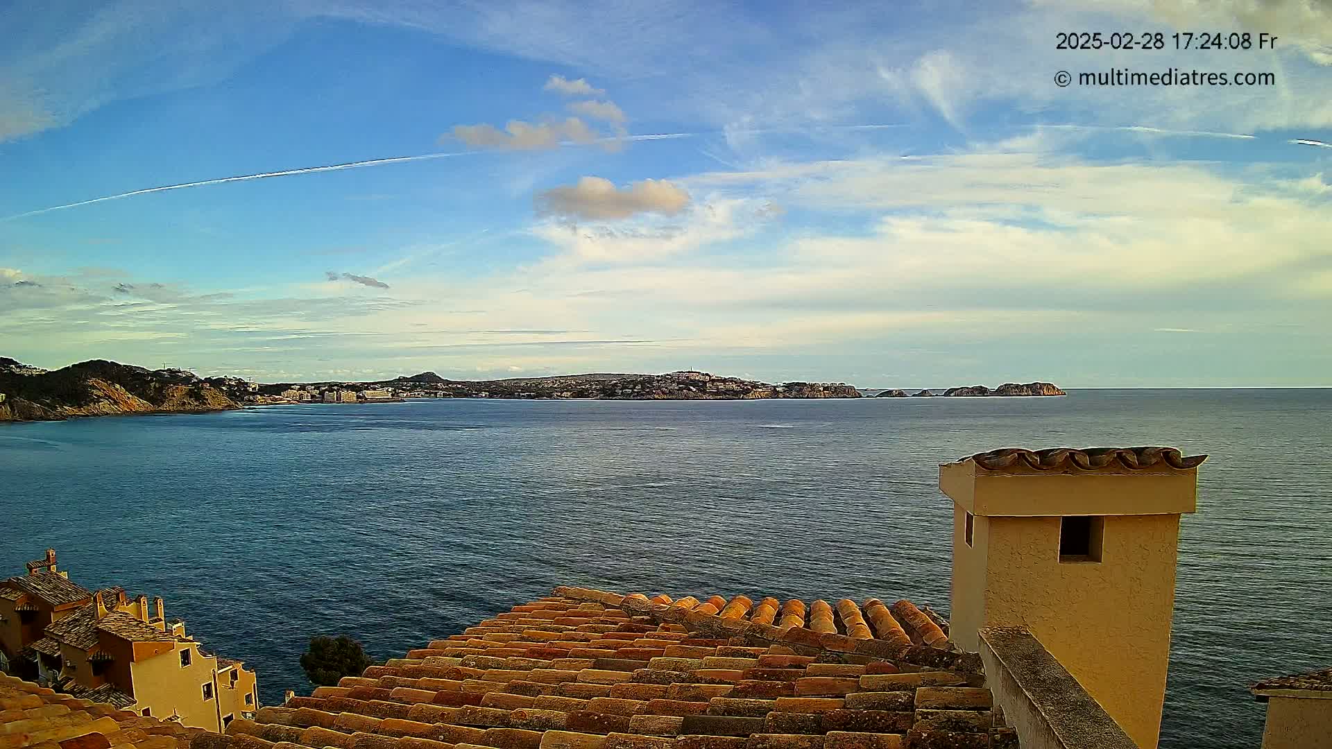 A partly cloudy sky overlooks a calm sea, with a distant coastline and buildings visible; a terracotta roof and chimney are in the foreground.