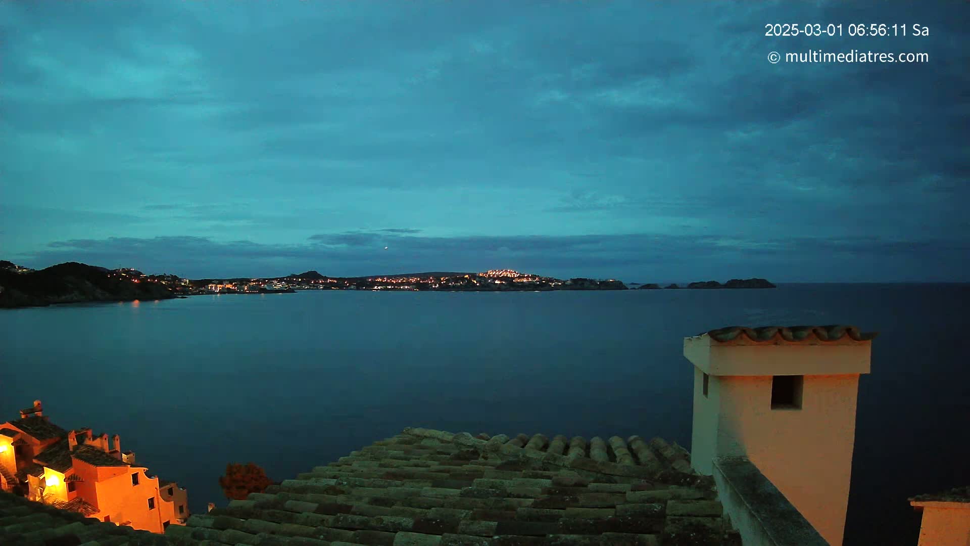 A twilight view of a calm, dark-blue sea with a town's lights visible across the water from a rooftop with terracotta tiles.