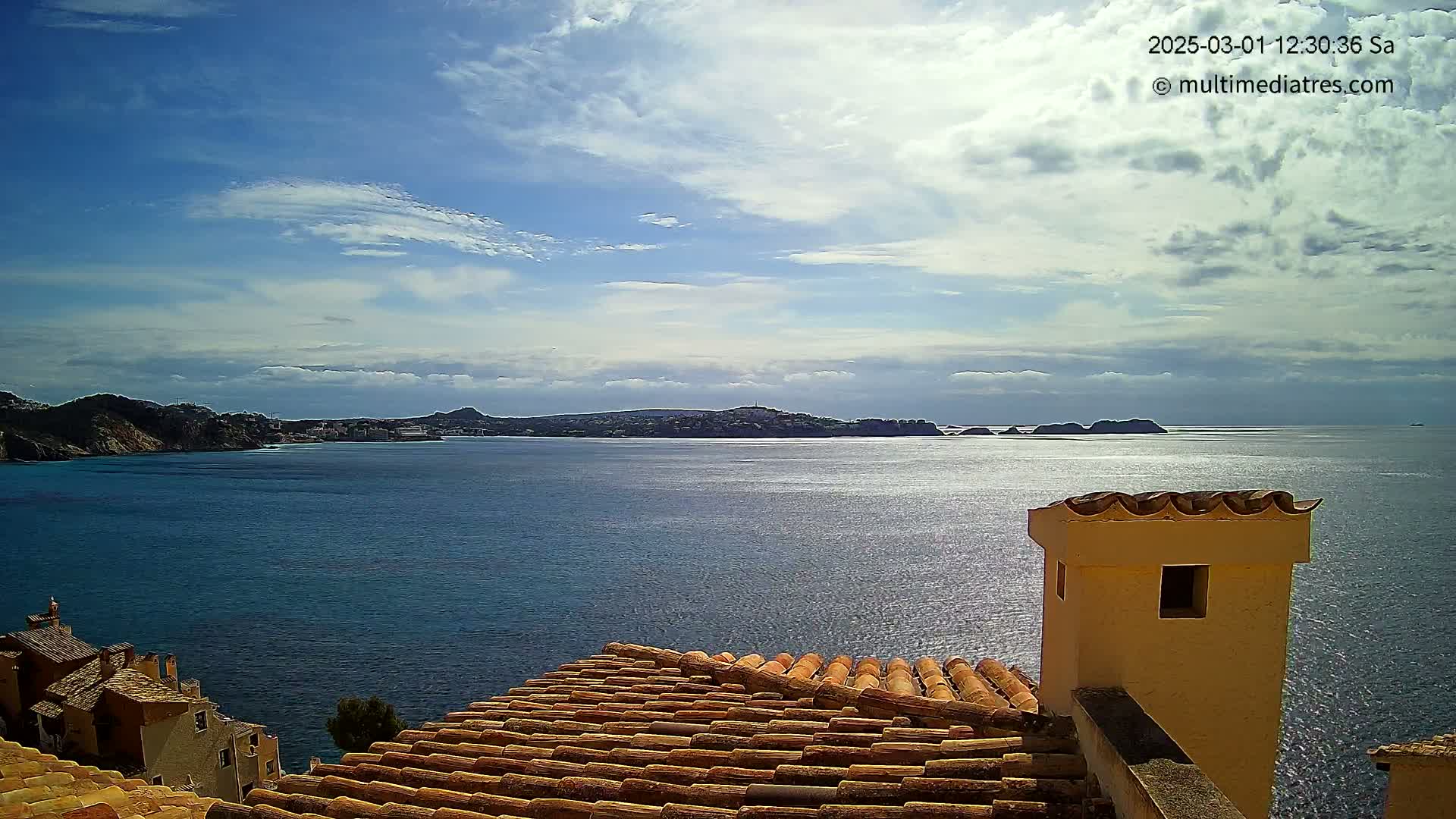 A partly cloudy sky overlooks a calm blue sea with distant hills and a portion of a tiled roof with a chimney in the foreground.