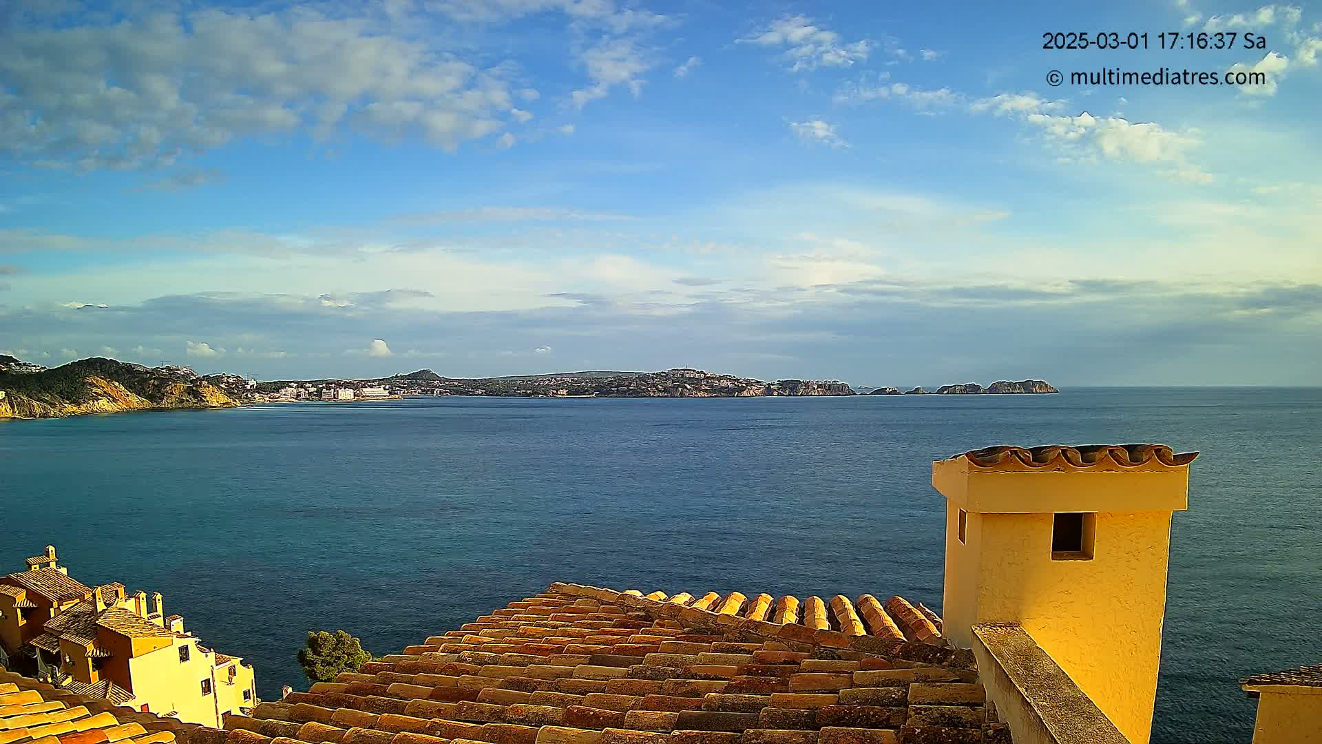 A partly cloudy sky overlooks a calm ocean with landmasses in the distance, viewed from a rooftop with terracotta tiles and a yellow chimney.