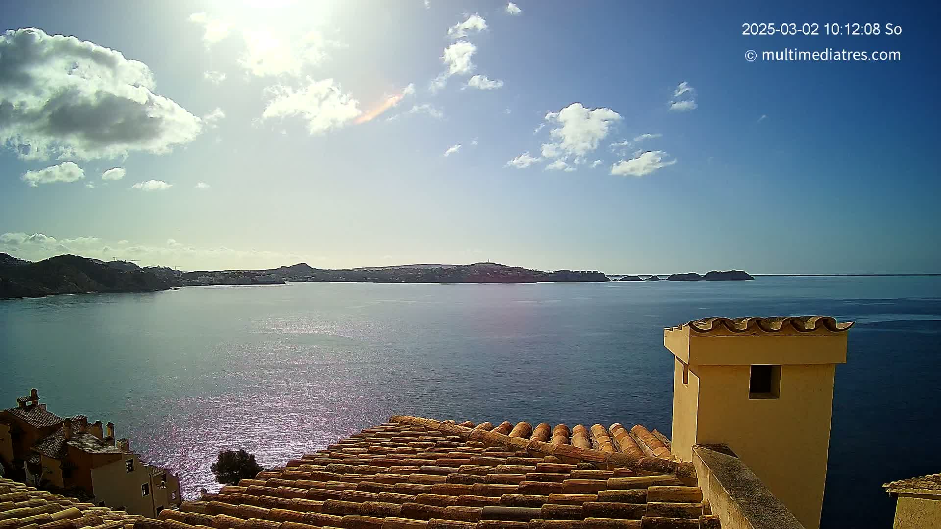 A sunny day with some clouds shows a view of a calm blue sea, with distant hills and islands, seen from a rooftop with terracotta tiles and a short chimney.