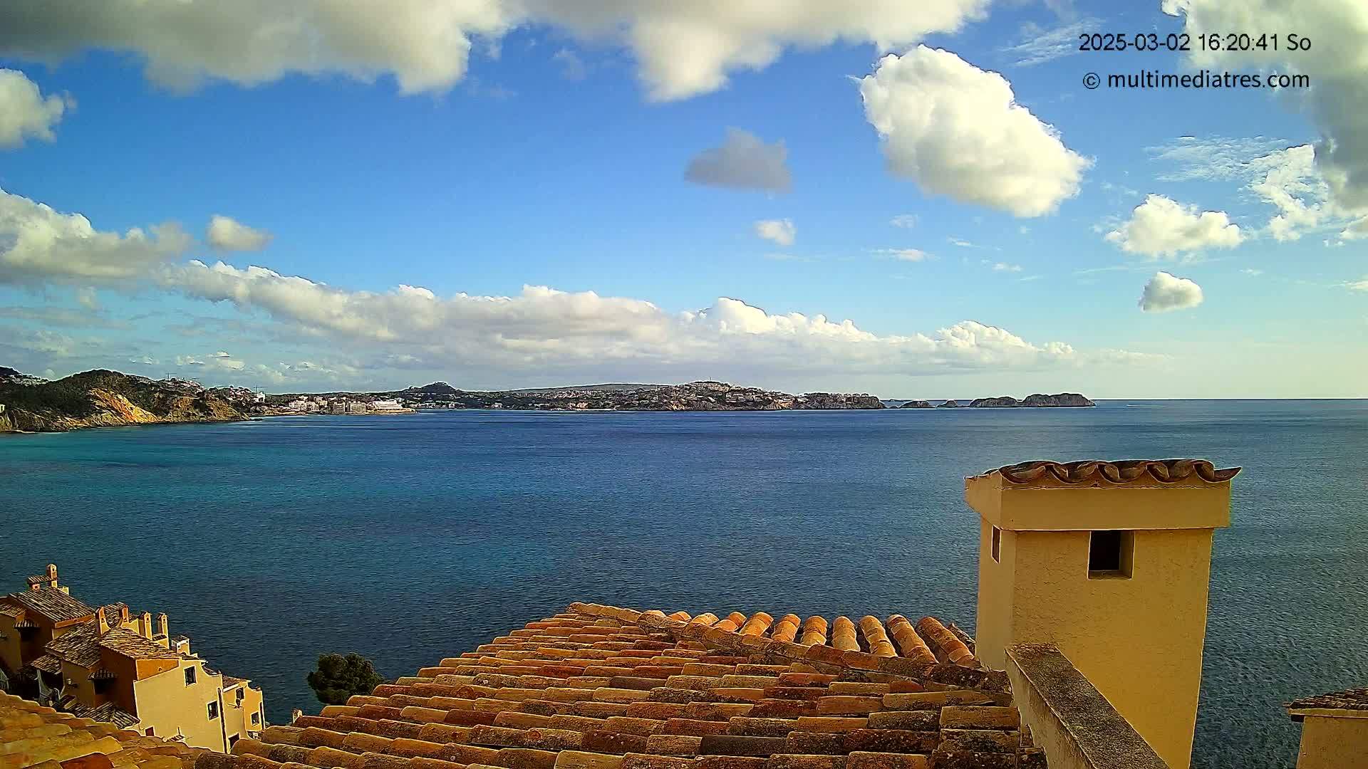 A partly cloudy blue sky overlooks a calm, teal sea with a distant coastline, viewed from a tiled rooftop with a tan chimney.