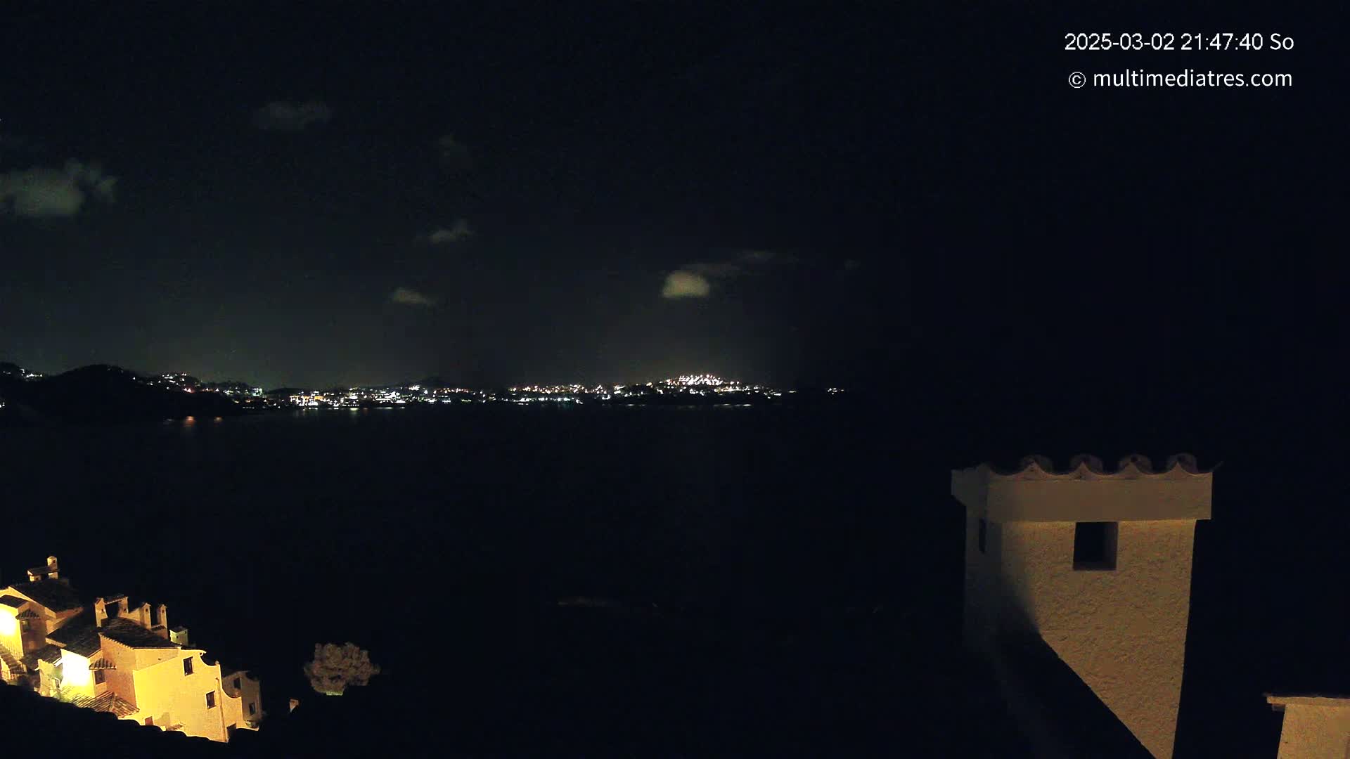 A nighttime view of a city skyline across a dark body of water, with a few clouds in the night sky, and a portion of a light-colored building in the foreground.