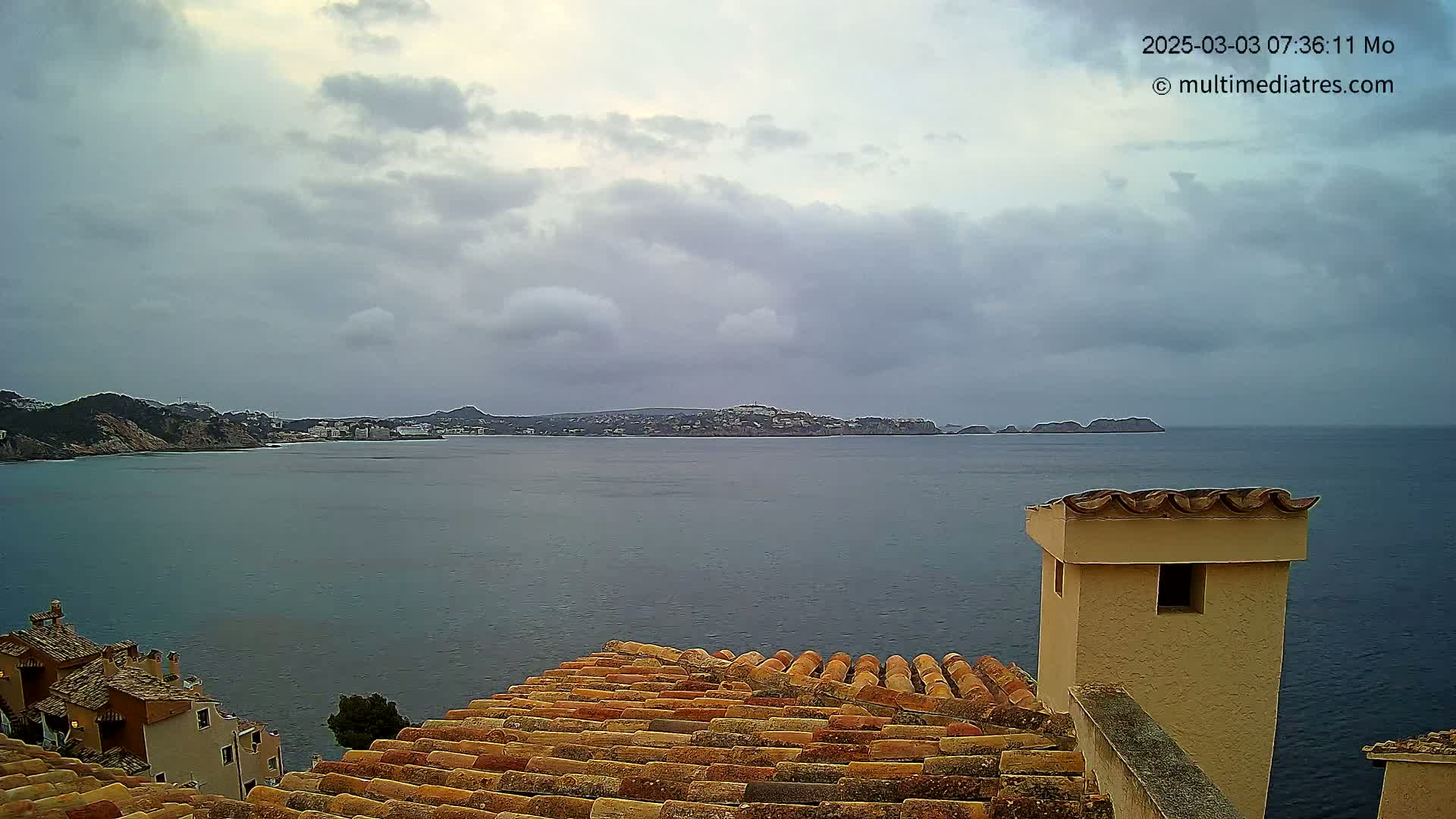 A view of a calm sea with distant land under a cloudy sky, seen from a building with a terracotta roof and a tan chimney.