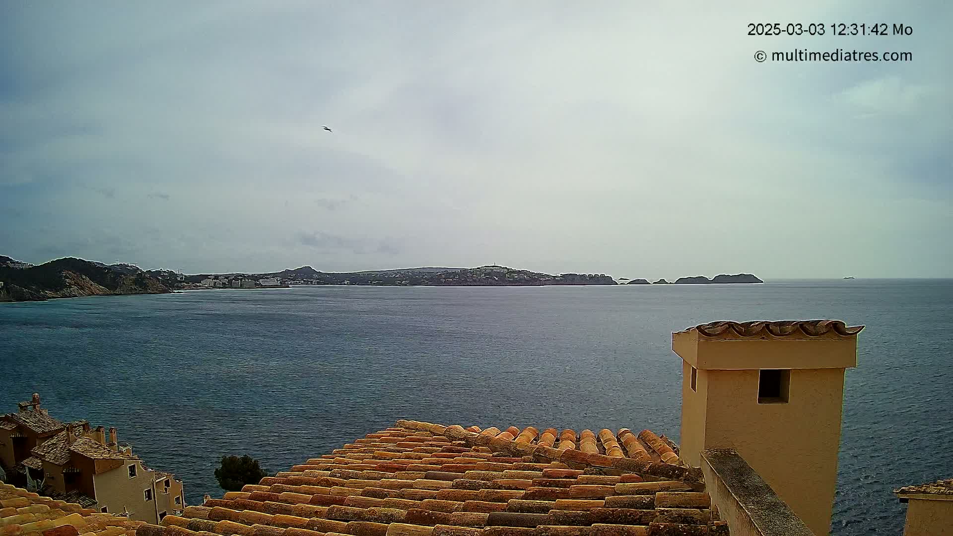 A terracotta roof overlooks a calm, blue sea with a distant coastline under a cloudy sky.