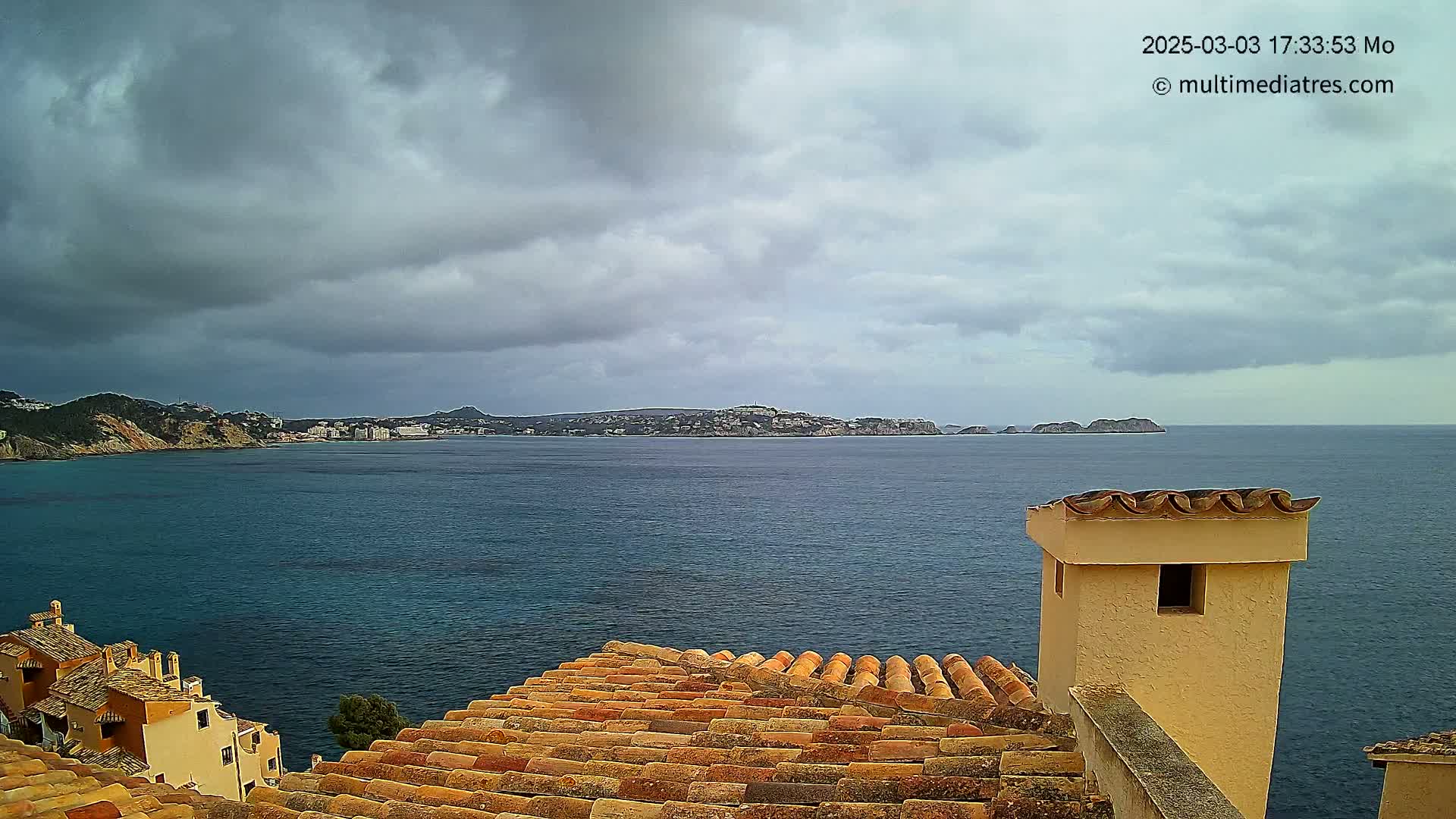 A view from a terracotta roof shows a calm sea under a cloudy sky, with a distant coastline visible.