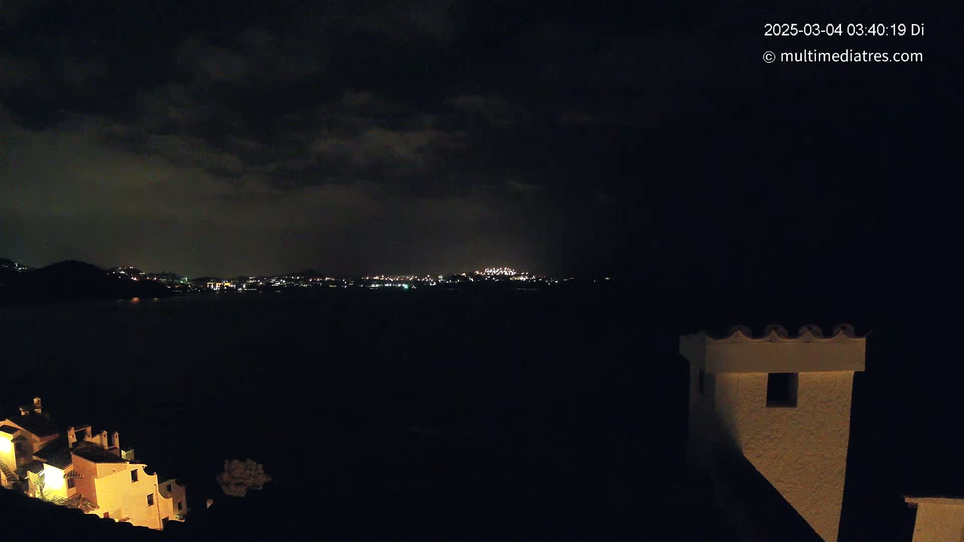 A nighttime coastal scene under a cloudy sky shows a distant city illuminated with lights across a dark body of water, with a portion of a building and a chimney in the foreground.