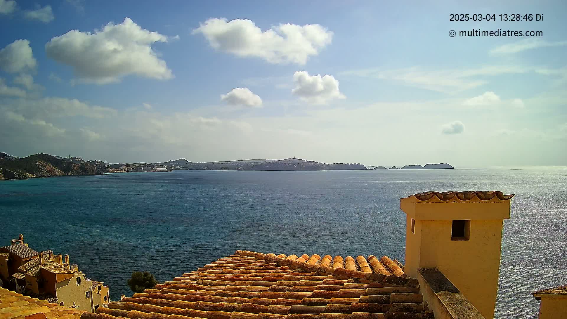 A partly cloudy sky overlooks a calm blue sea, with a distant island and a terracotta roof in the foreground.