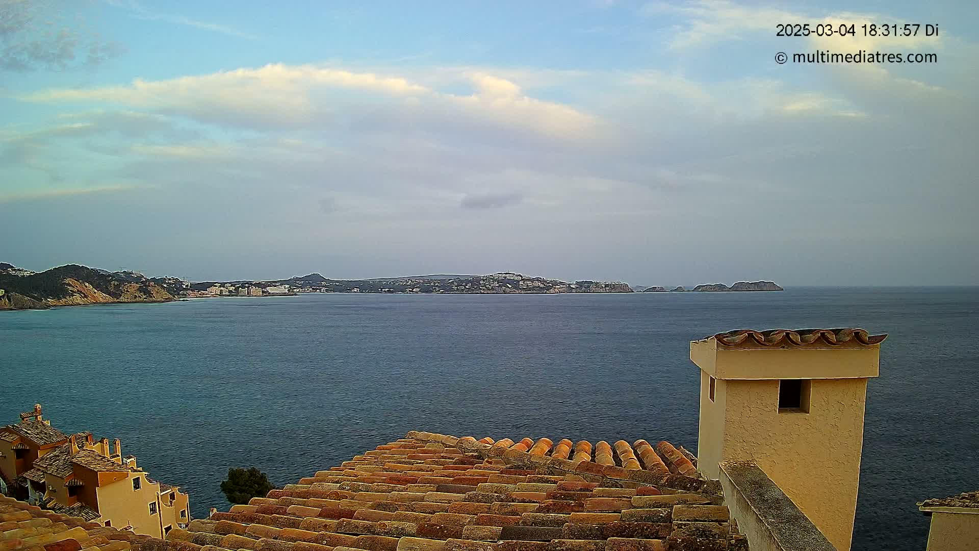 A coastal view shows a calm sea under a partly cloudy sky, with terracotta rooftops in the foreground and a distant shoreline.