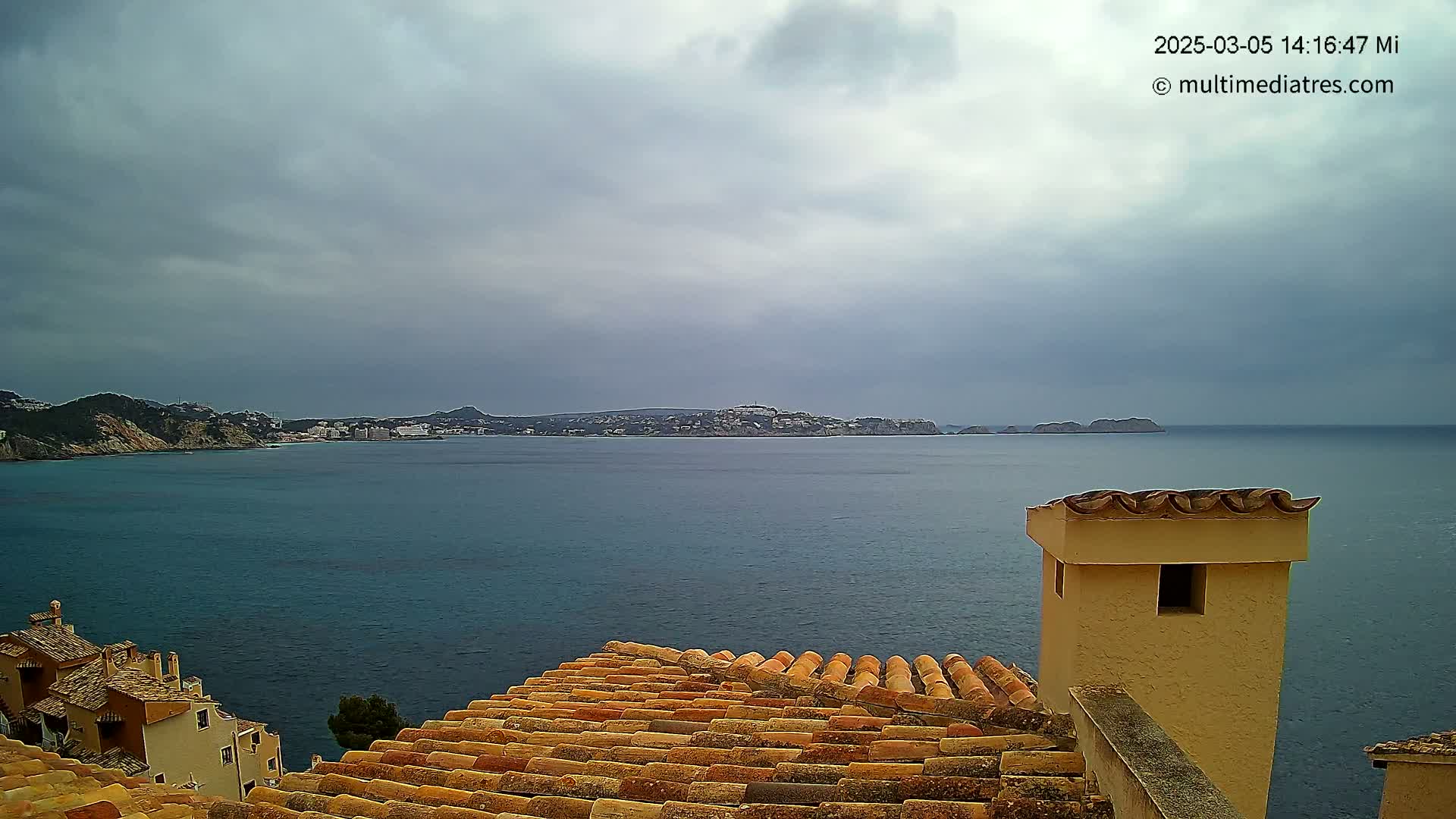 A coastal view under an overcast sky shows a calm sea separating a terracotta-roofed building from a distant town nestled against a mountainous shoreline.