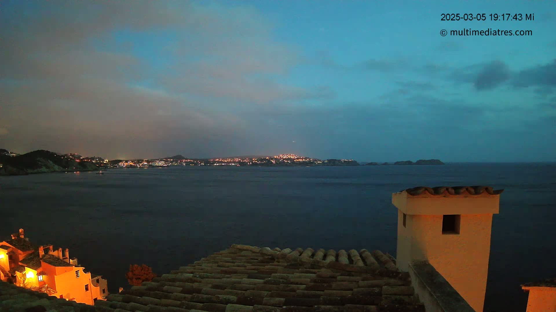 A nighttime view from a rooftop shows a calm body of water with a lit-up town on the far shore under a mostly clear, dusky blue sky.