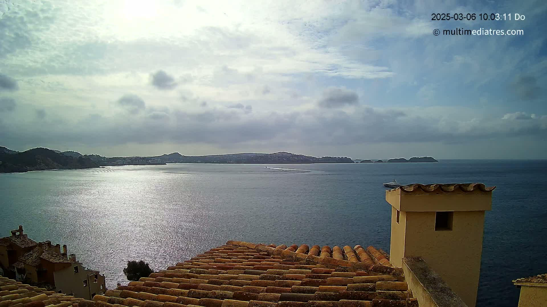A partly cloudy day overlooks a calm sea with distant hills and islands, viewed from a tiled rooftop.