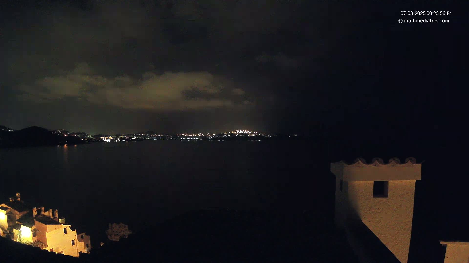 A nighttime view shows a dark body of water separating a dimly lit shoreline with buildings from a faraway city skyline under a cloudy sky.