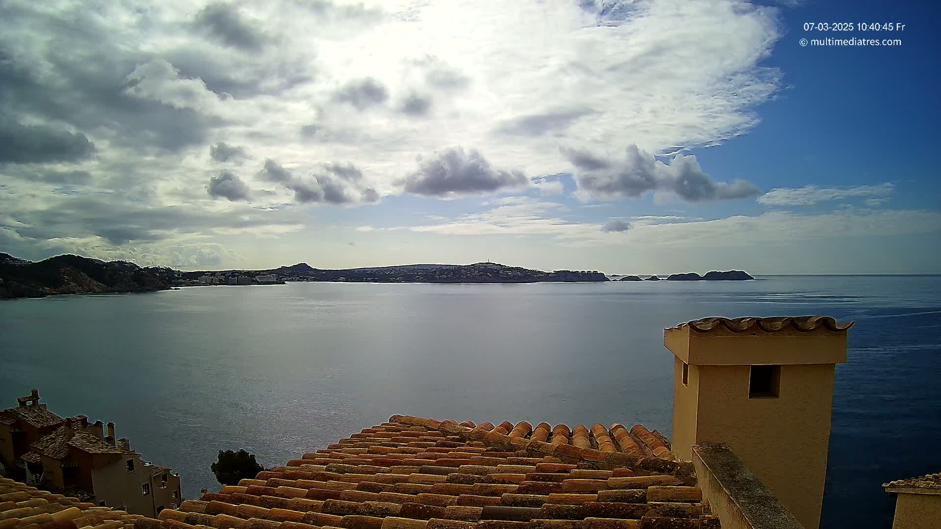 A partly cloudy sky overlooks a calm sea with distant land and buildings, viewed from a tiled rooftop with a chimney.