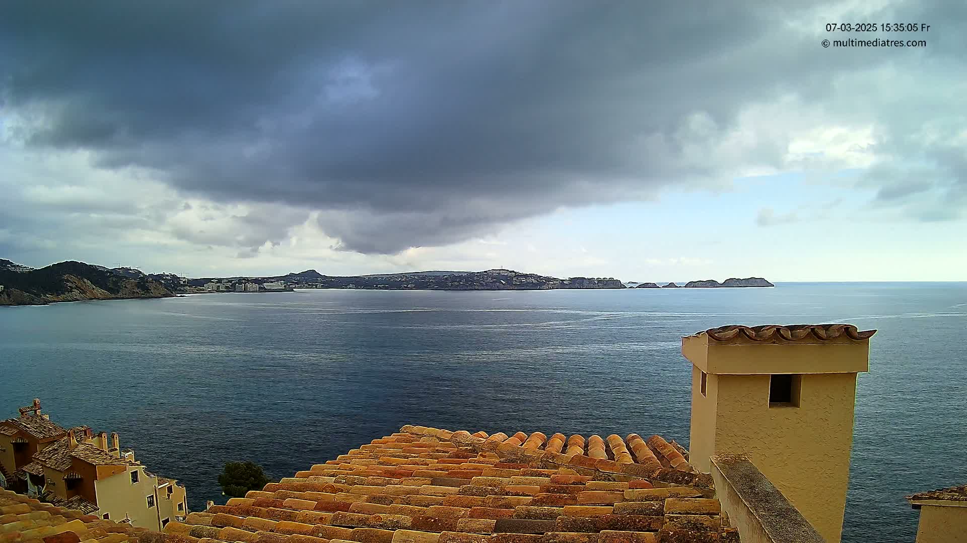 Overlooking a calm sea from a tiled roof, a coastal town is visible across the water under a partly cloudy sky.
