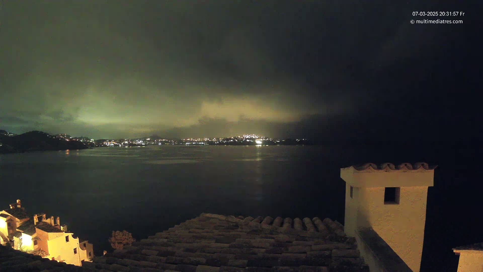 A nighttime view from a rooftop shows a calm body of water reflecting the lights of a city across the bay under a cloudy, dark sky.