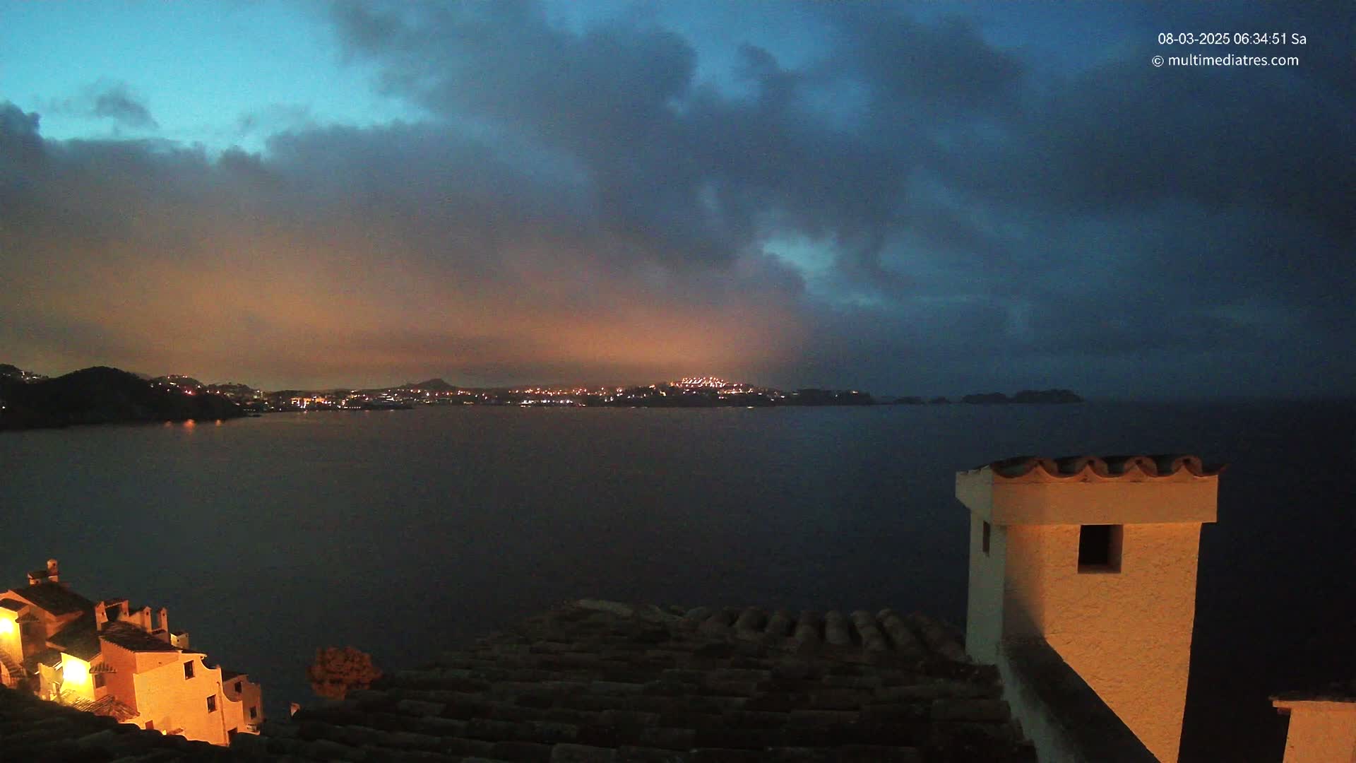 A nighttime coastal scene shows a dark, cloudy sky over a calm body of water with a city's lights visible on the far shore, and a portion of a building with a tiled roof in the foreground.