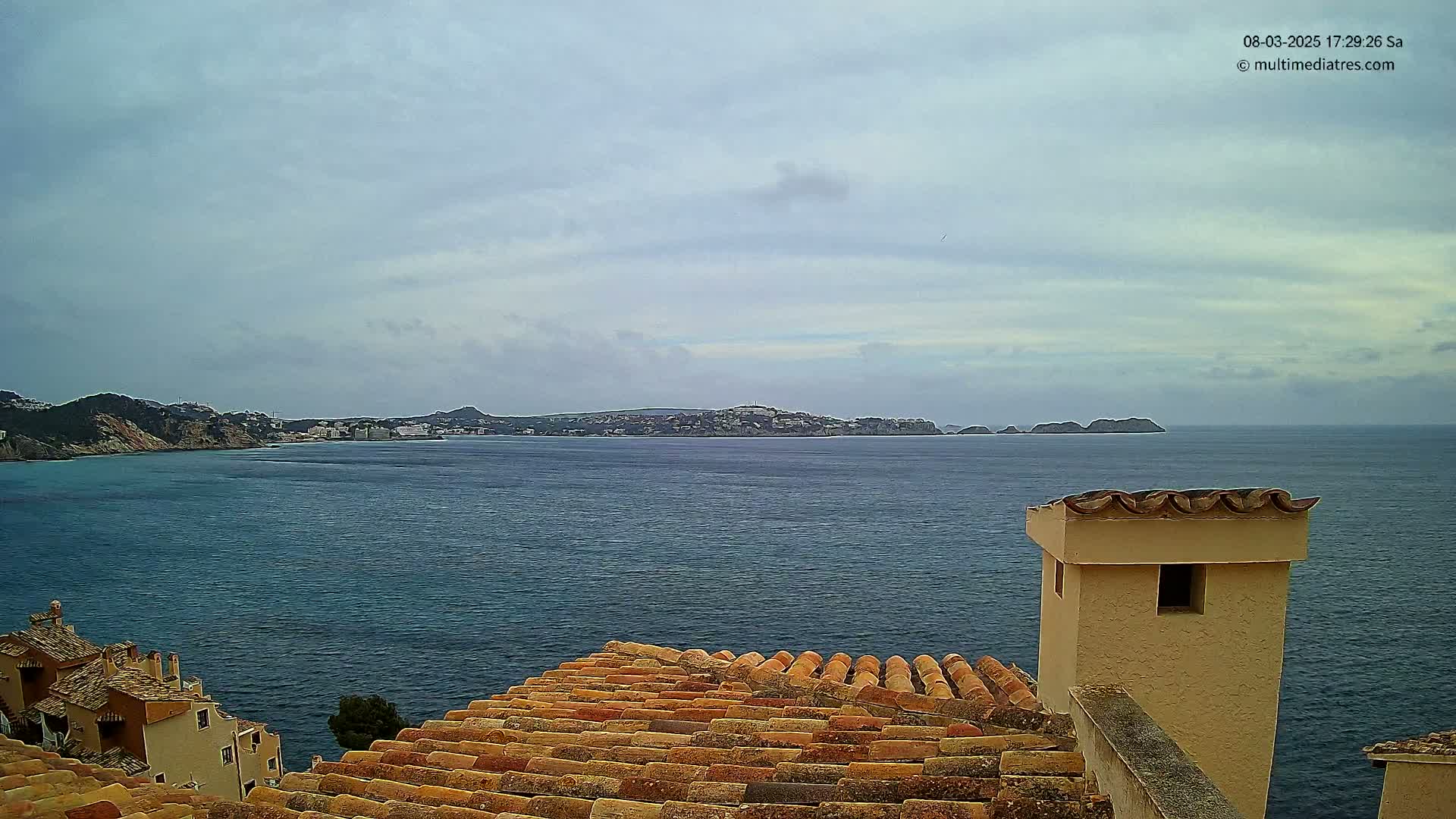 A coastal view under a cloudy sky shows a calm sea with a distant town visible across the water, partially obscured by a closer coastline, as seen from atop a building with a terracotta roof and a tan chimney.